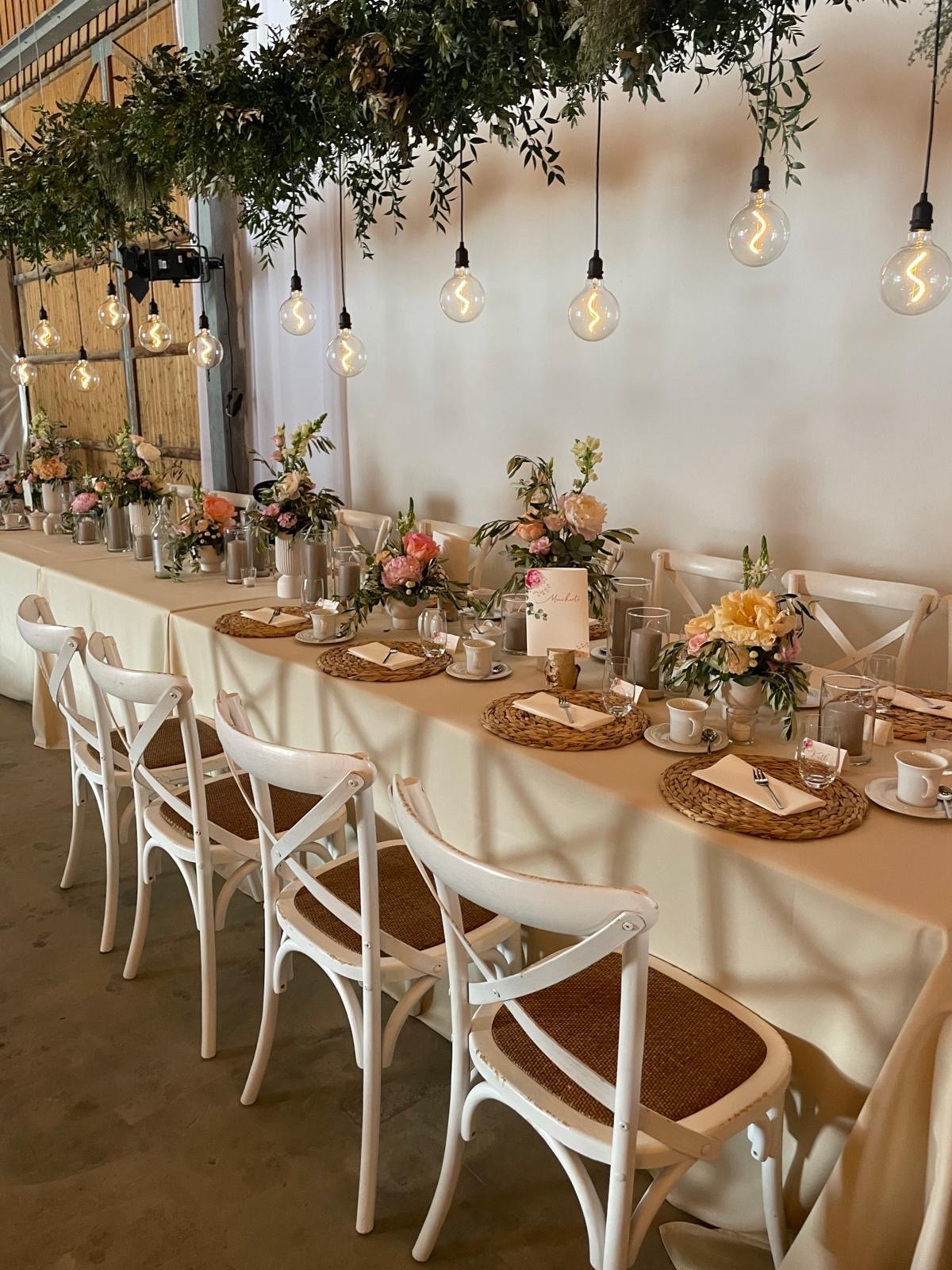 Elegante Hochzeitstischdekoration mit weißen Stühlen, beige Tischdecke, Blümengestecken, Tellern, Tassen, und hängenden Glühbirnen; über dem Tisch schwebende Pflanzenarrangements.