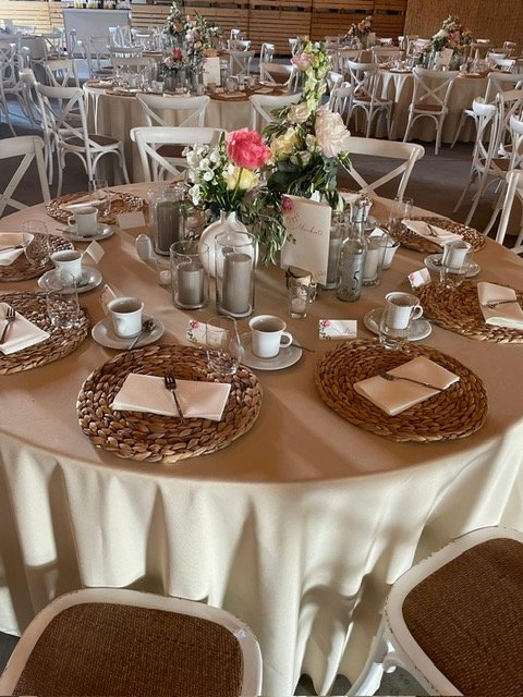Rund gedeckter Tisch für eine Veranstaltung mit weißen Tischdecken, geflochtenen Tischsets, Gedeck aus Kaffeetassen und Tellern, sowie einem Blumenarrangement in der Mitte.