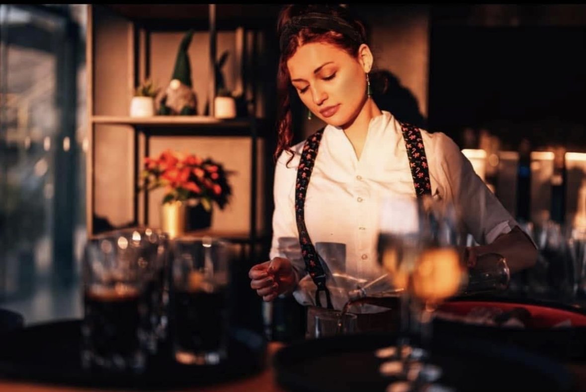 Eine Frau in einem Café oder Restaurant, die ein Getränk zubereitet, vor einem Regal mit Pflanzen und Dekorationen.