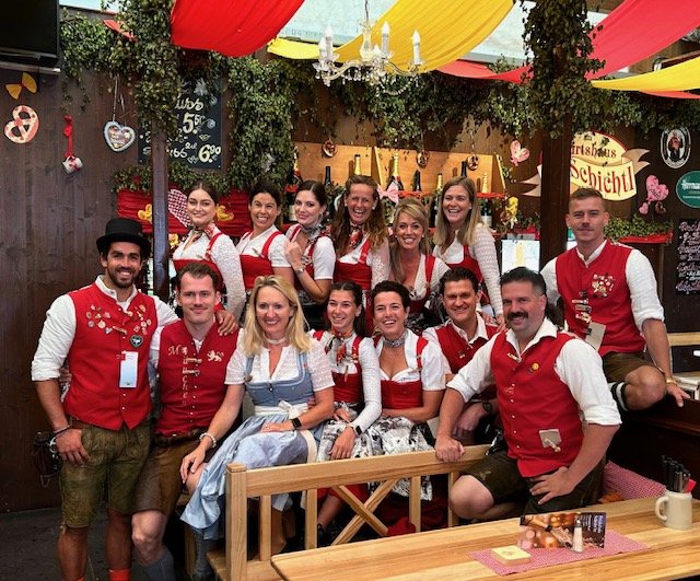 Gruppe von Menschen in traditioneller bayerischer Tracht, in einem festlich dekorierten Raum mit Oktoberfest-Thema.