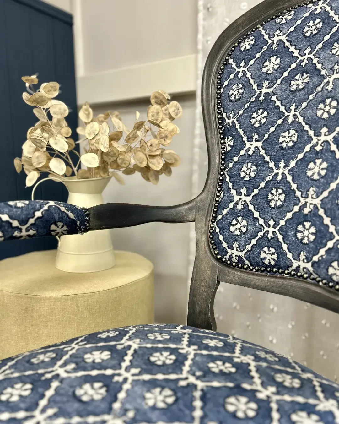 Close-up of a vintage chair with a blue and white patterned upholstery, featuring floral and geometric motifs, with a wooden armrest. In the background, a cream-colored vase with dried flowers and a beige ottoman are visible, against a white wall and a dark blue cabinet.