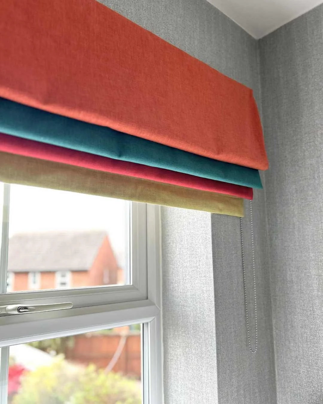 Close-up of colorful roman blinds in orange, teal, pink, and yellow above a window with a view of a house and garden