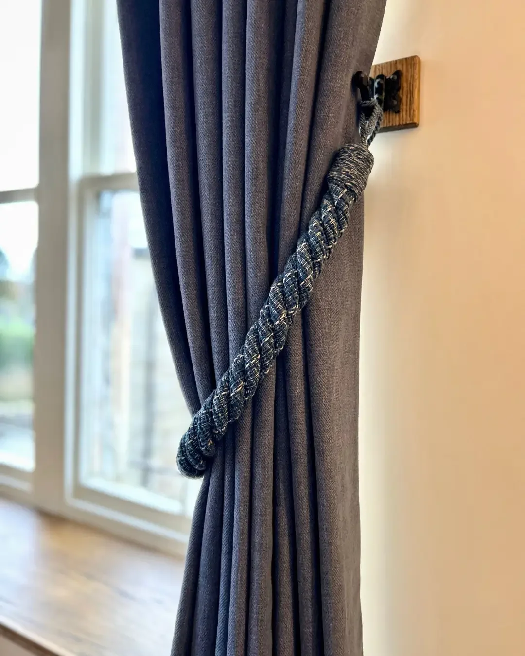A close-up of a dark blue curtain pulled back and held with a braided blue Laura Ashley curtain tie, with a window in the background.