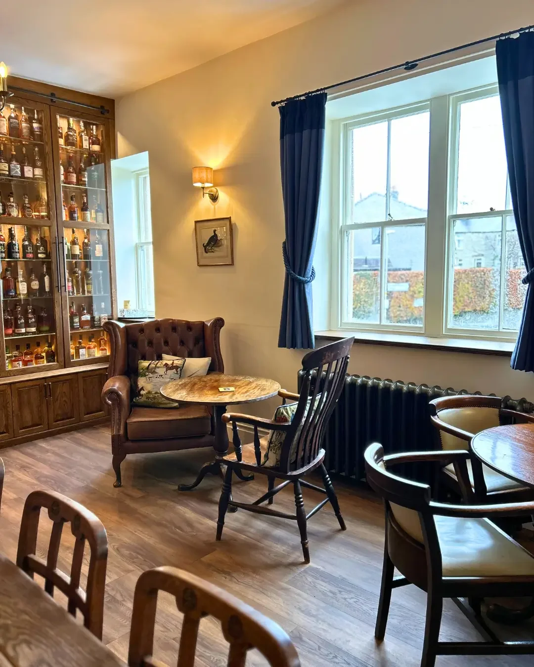 Cosy pub interior with wooden flooring, large window with blue curtains, a wooden cabinet filled with whiskey, vintage chairs, leather armchair with handmade cushions small round wooden table.