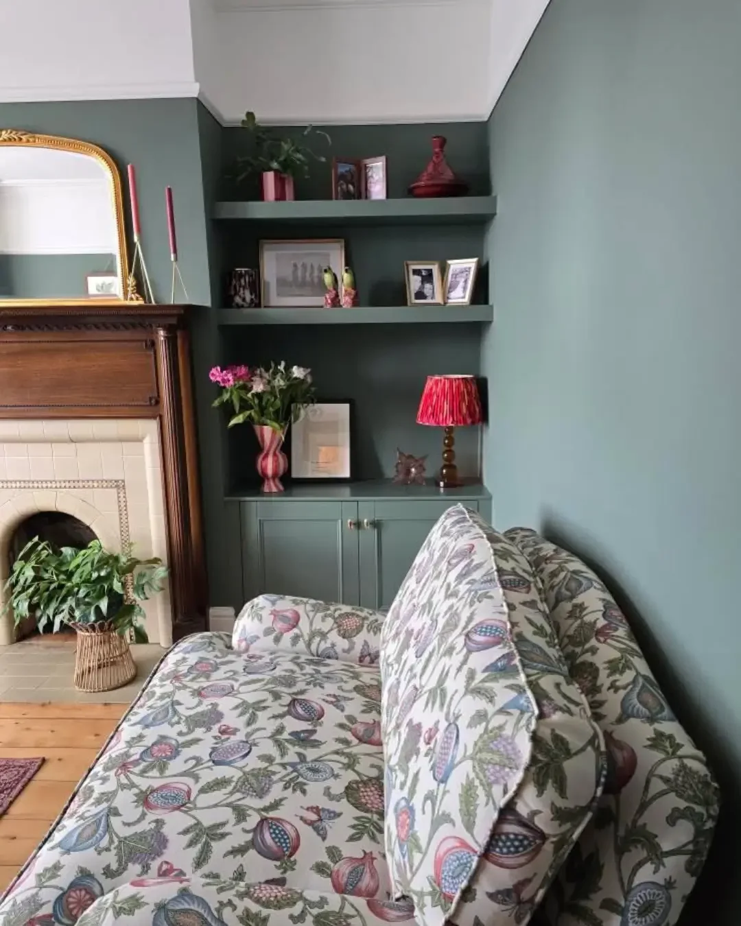 A cozy living room corner featuring a floral-patterned sofa, a green cabinet with decorative items and a red table lamp, a bookshelf with framed photos and plants, and a fireplace with a potted plant in front.