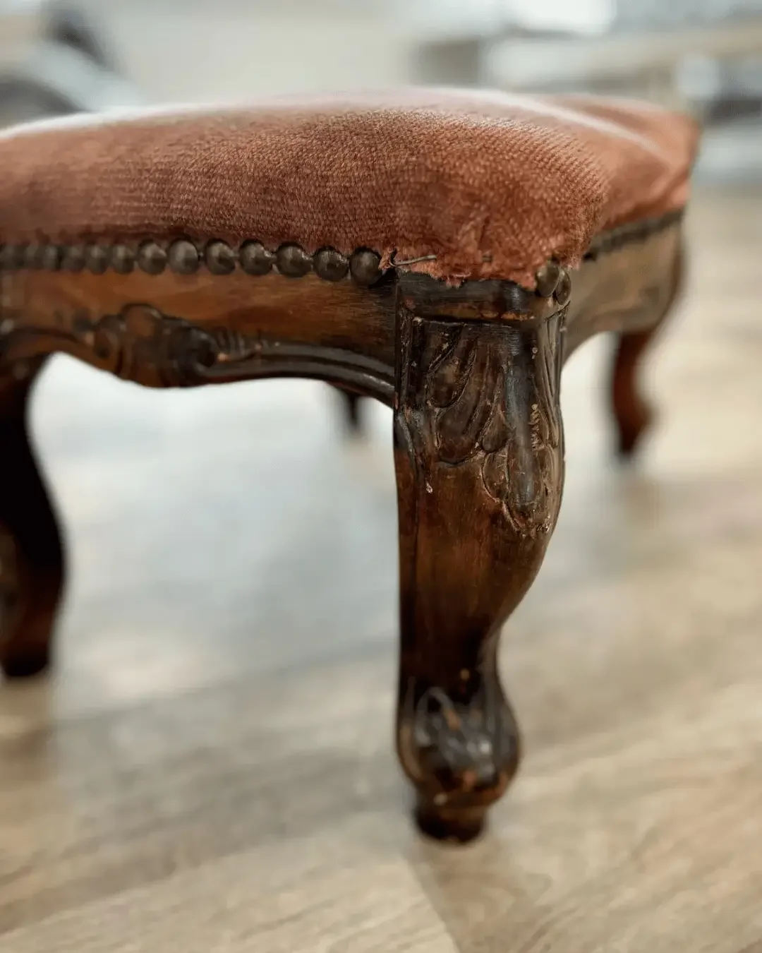 Close-up of a vintage wooden footstool with worn, reddish velvet fabric upholstery and decorative carved leg details. The before shot of one of our reupholstery transformations.