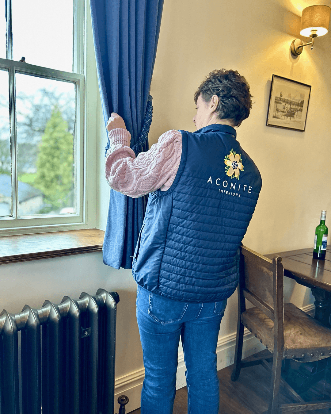 Alison from Aconite Interiors hanging the bespoke commerical curtains in the Lower Buck Pub Waddington. She is wearing a pink sweater and a navy blue gilet with the text "ACONITE INTERIORS" and logo on the back.