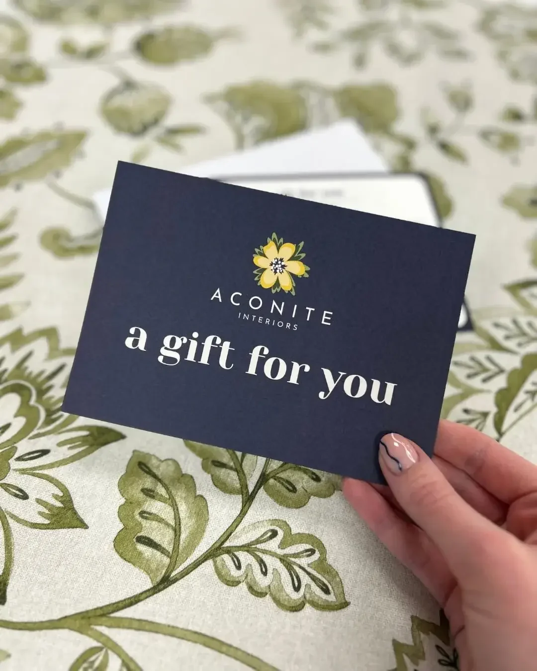 Hand holding an Aconite Interiors Gift Voucher with green and white botanical print The Pure Edit fabric in the background