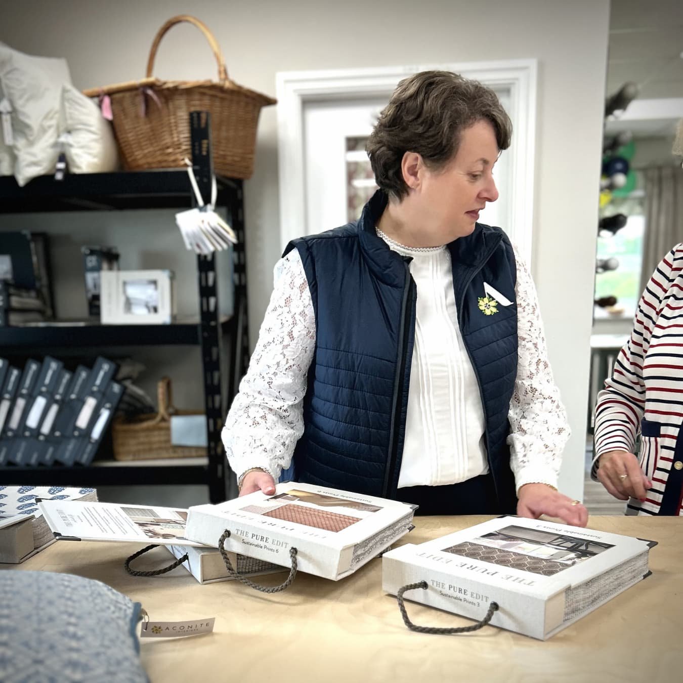 Alison holding a design consultation with a customer. She is looking through fabric sample books by The Pure Edit. Alison is wearing a white blouse and an Aconite Interiors branded gilet
