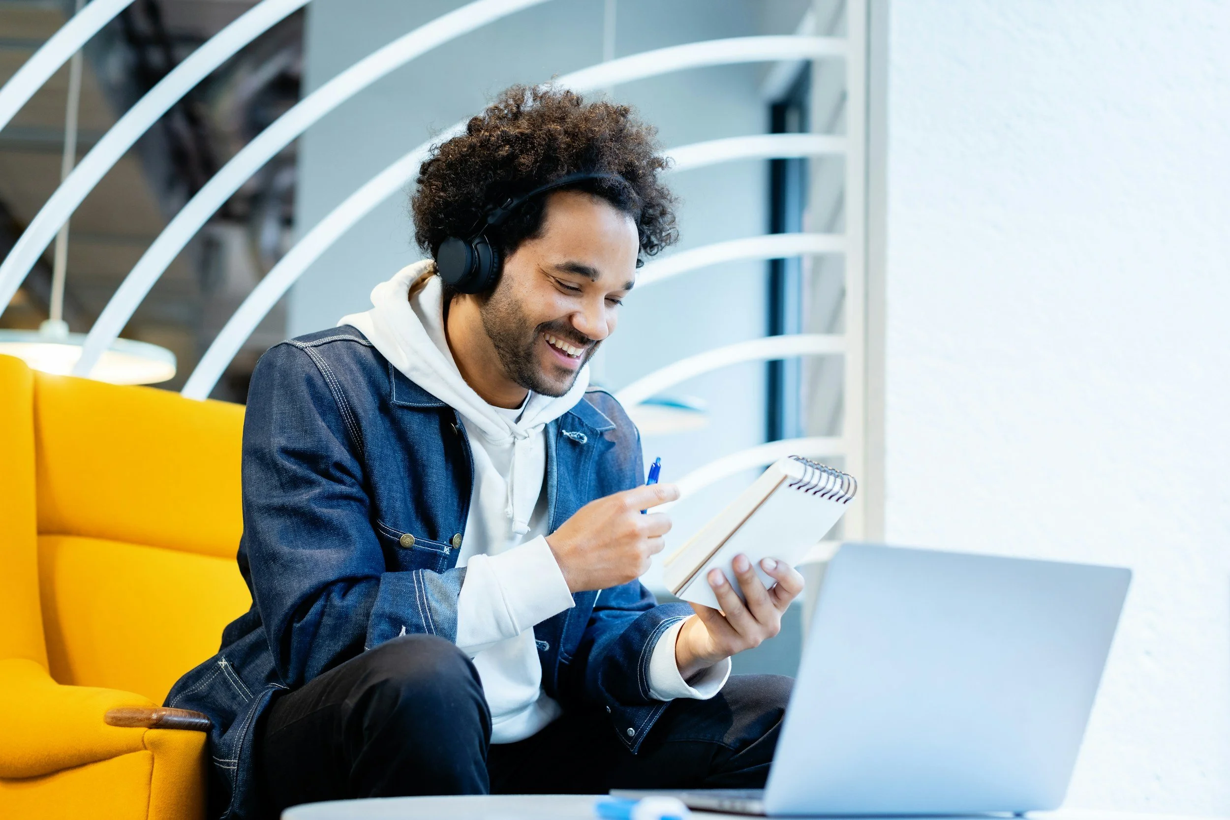 Person wearing headphones smiling and writing in a notebook, seated in a bright yellow chair, using a laptop.