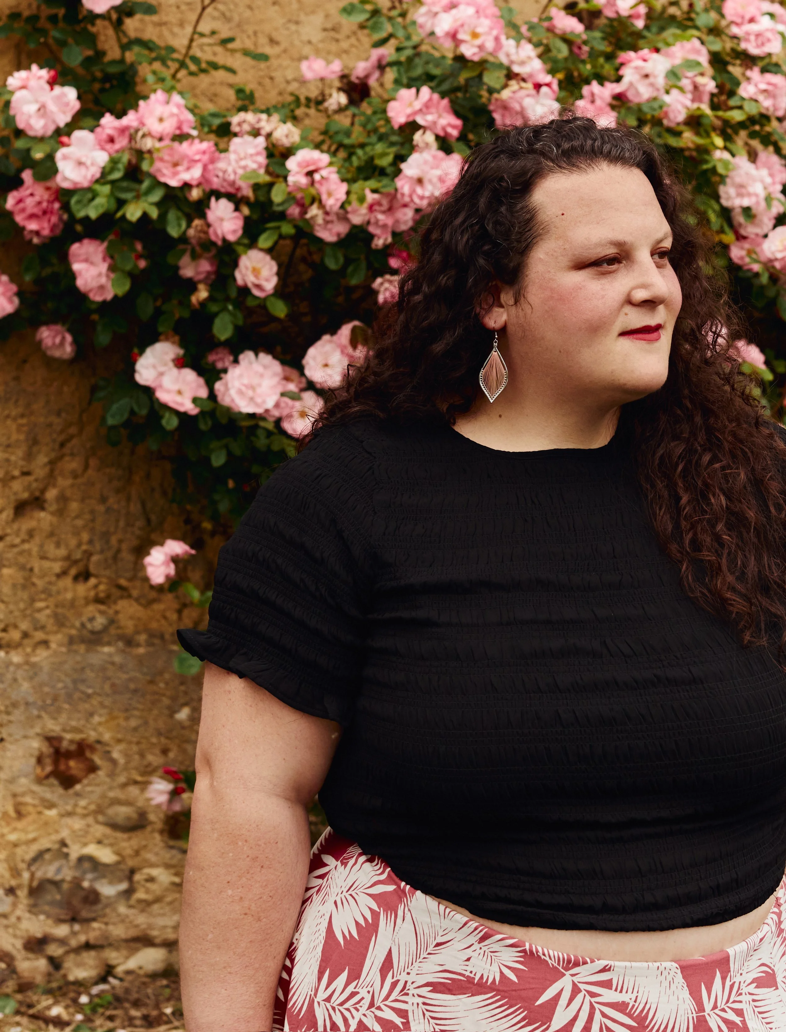 An embodied woman with long curly hair wearing a black top, pink and white leafy patterned skirt, and dangle earrings, standing outside near a bush with pink flowers.