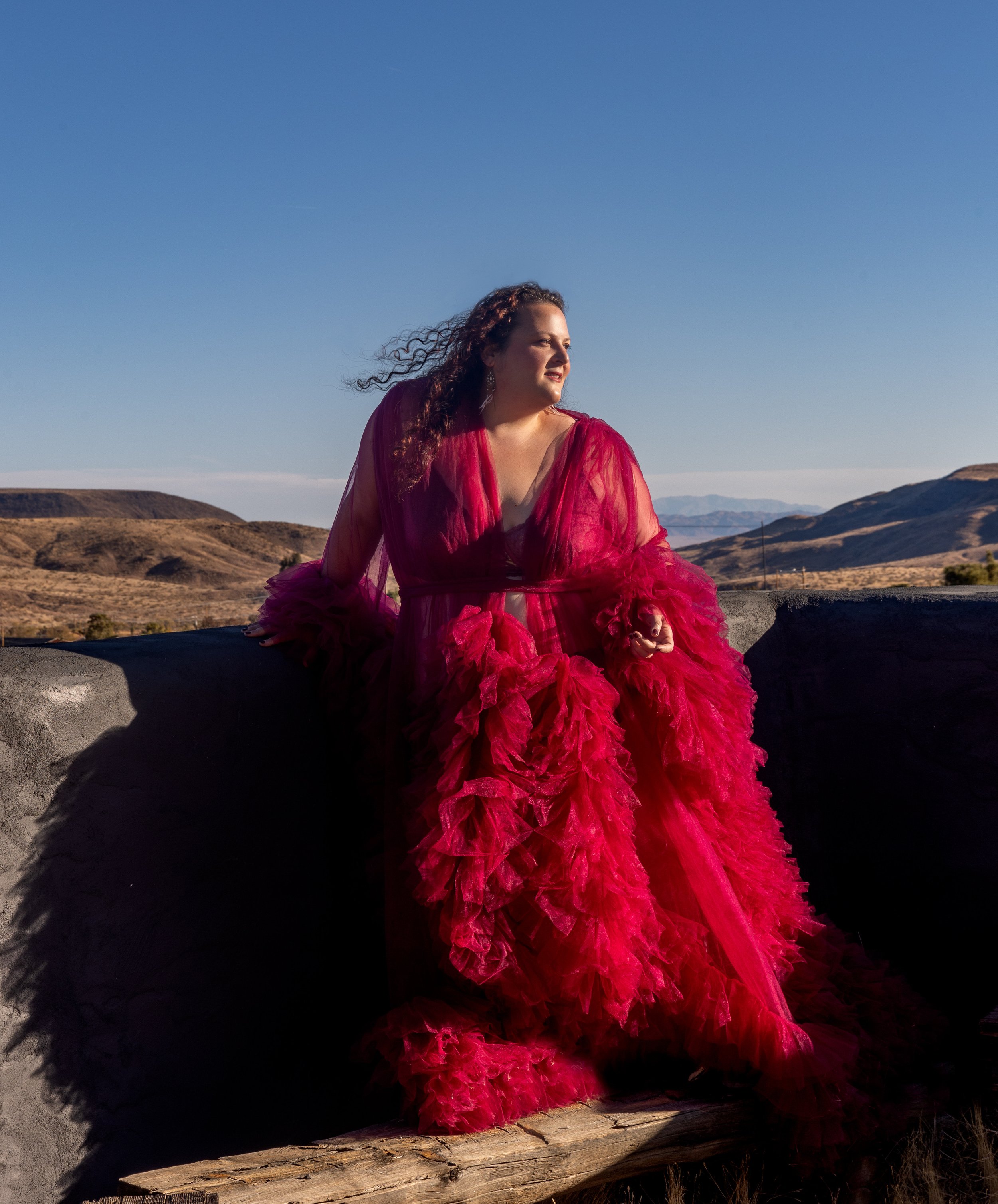 Embodied woman in a vibrant red, ruffled gown sitting outdoors on a log with a desert landscape and mountains in the background.