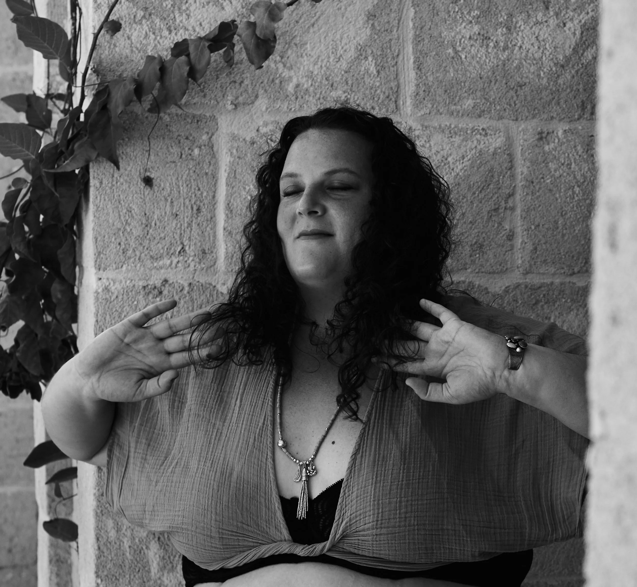 Black and white photo of an embodied woman with curly hair, eyes closed, making a relaxed facial expression, with hands raised near her shoulders in front of a brick wall and some leaves. She is adorned with a necklace.