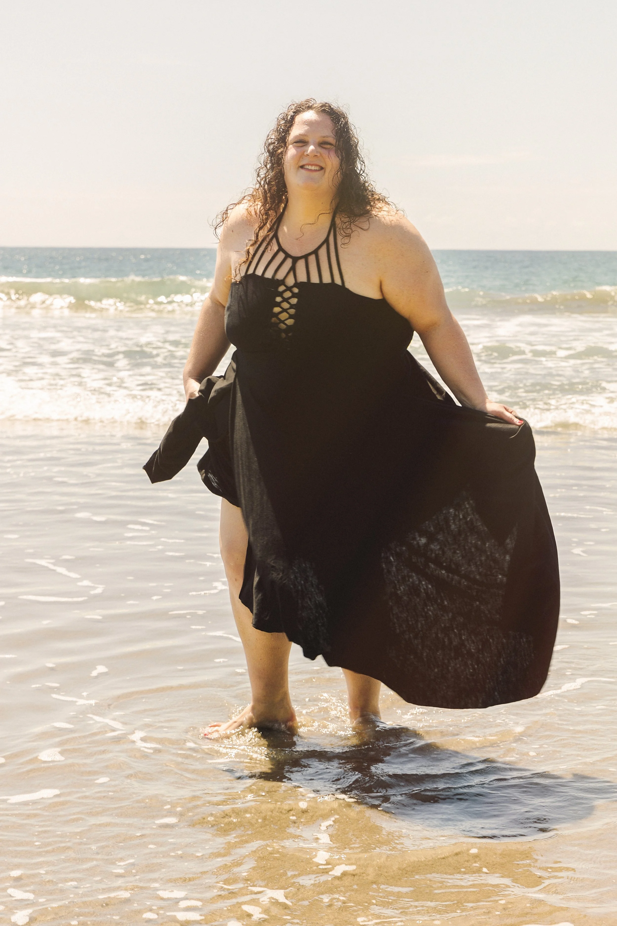 Embodied woman in a black dress standing in shallow ocean water at the beach with waves and sky in the background.