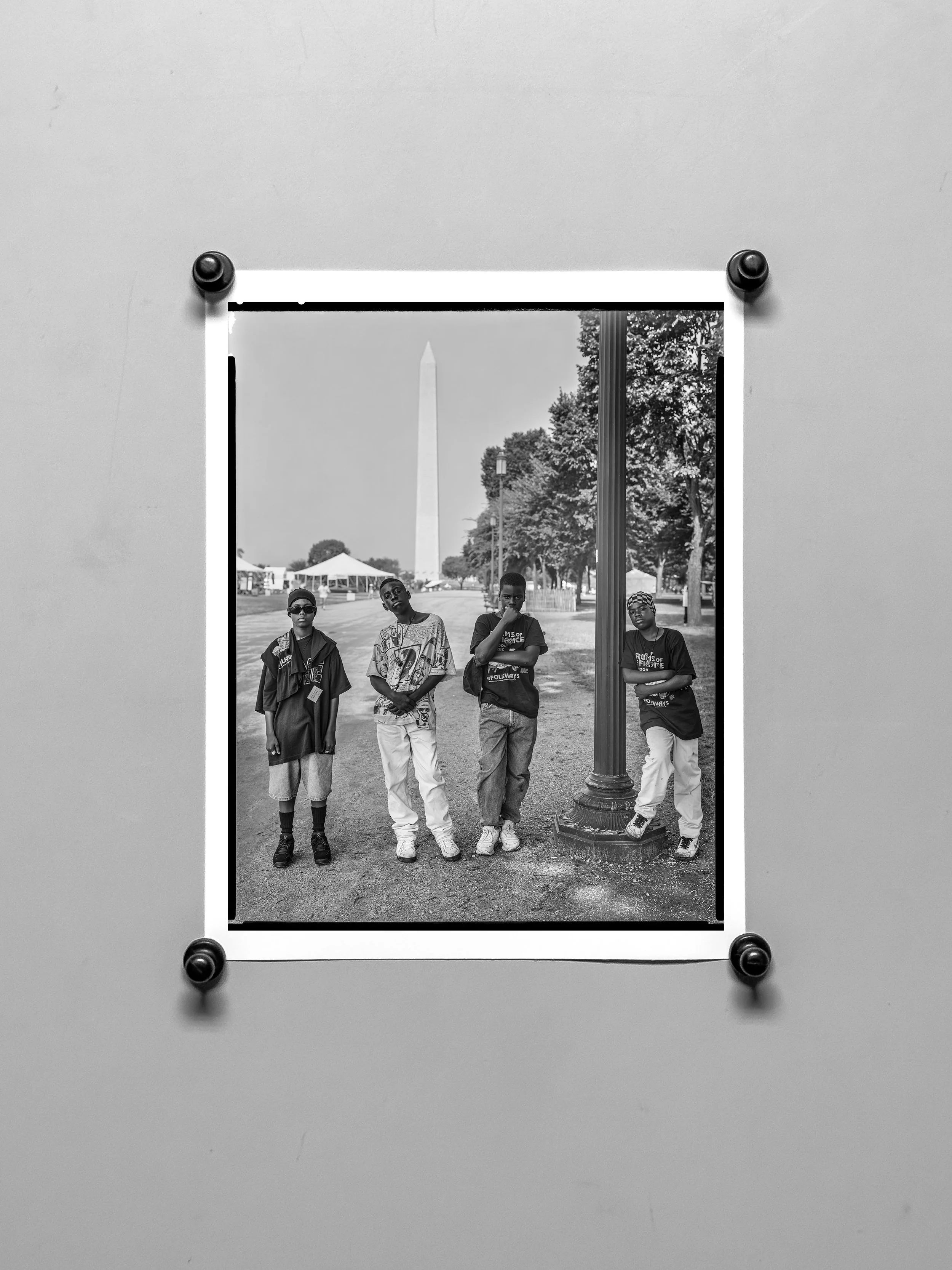 Black and white photograph of four young boys standing near a lamppost in a park, with trees and a distant Washington Monument in the background.