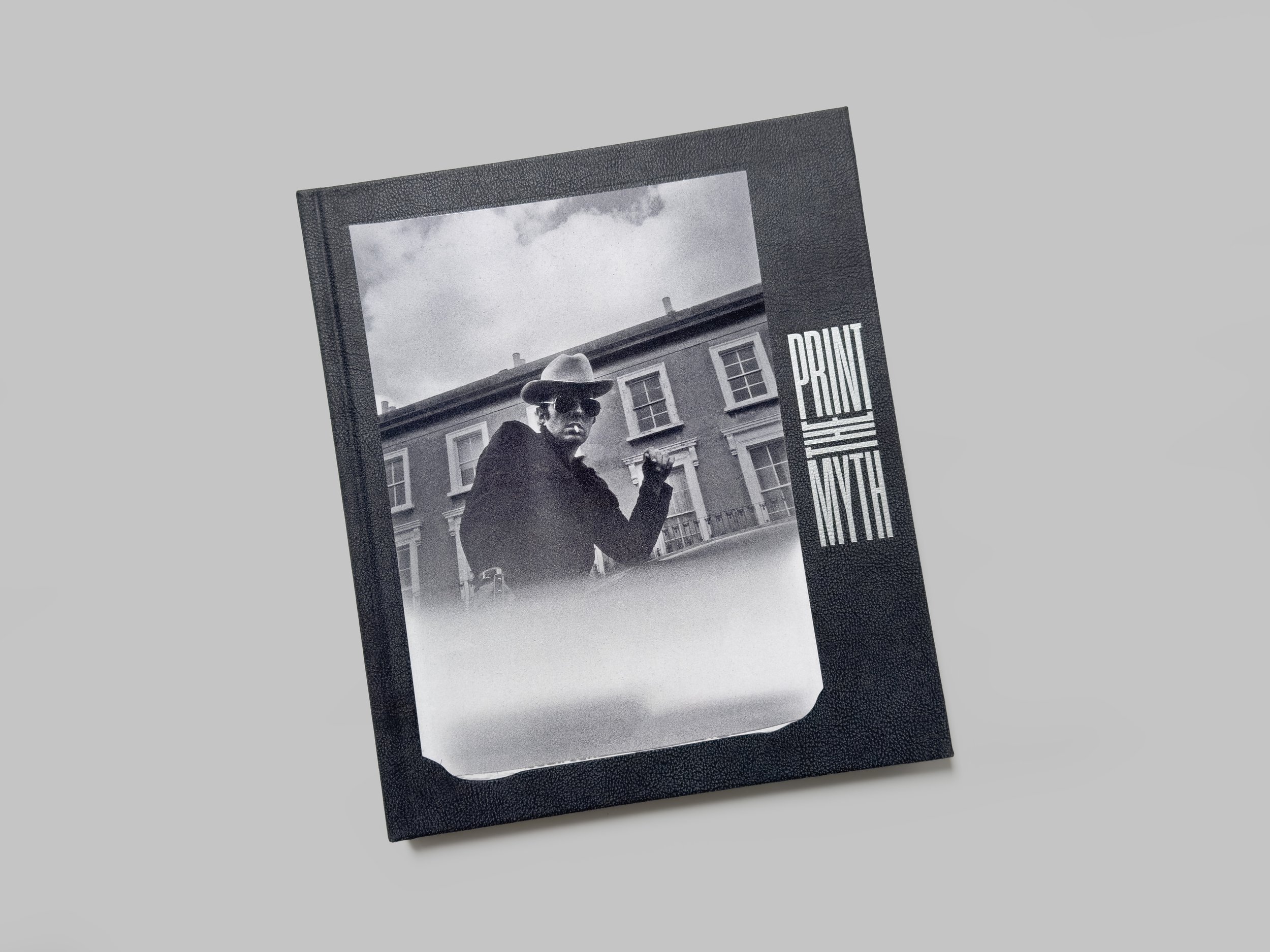 Black and white photograph of a person wearing sunglasses and a hat, standing in front of a building, inside a black photo frame with the words "PRINT WITH" on the side.