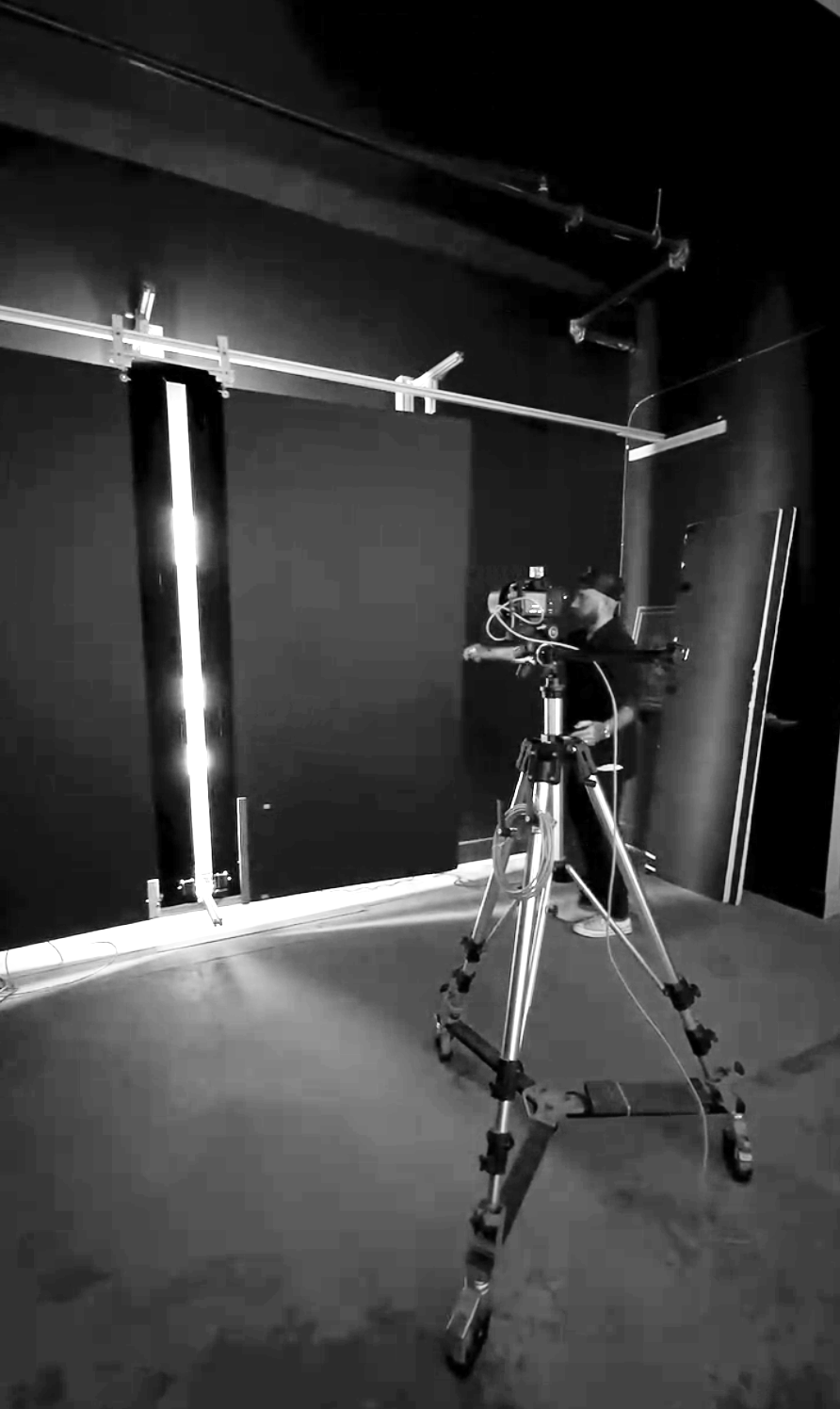 Black and white photo of a camera on a tripod set up in a dark room with a person in the background adjusting equipment near a black wall and metal rods.