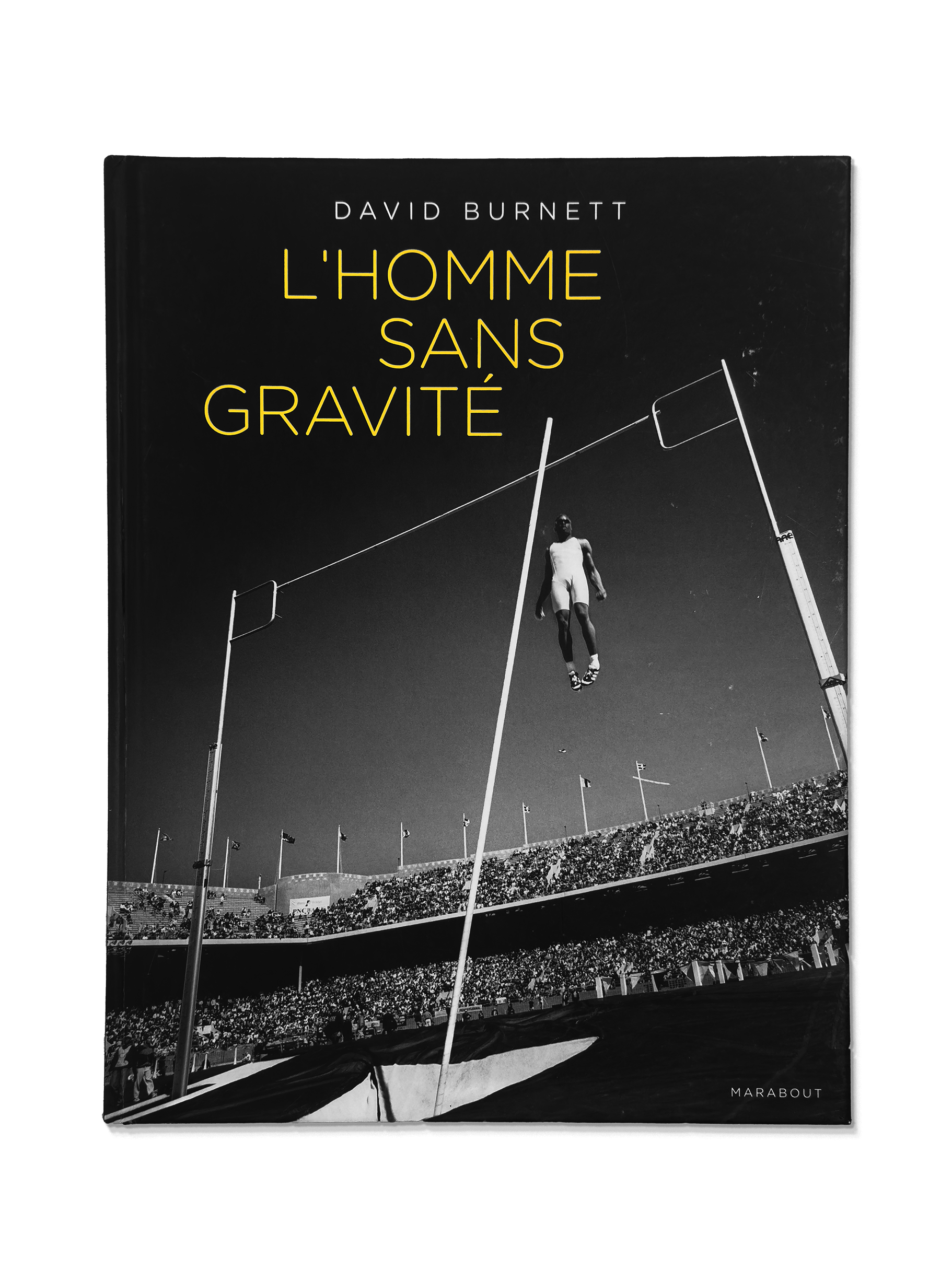 Book titled 'L'Homme Sans Gravité' by David Burnett features a black and white photograph of a high jump during a track and field event, with a crowd in the stadium.