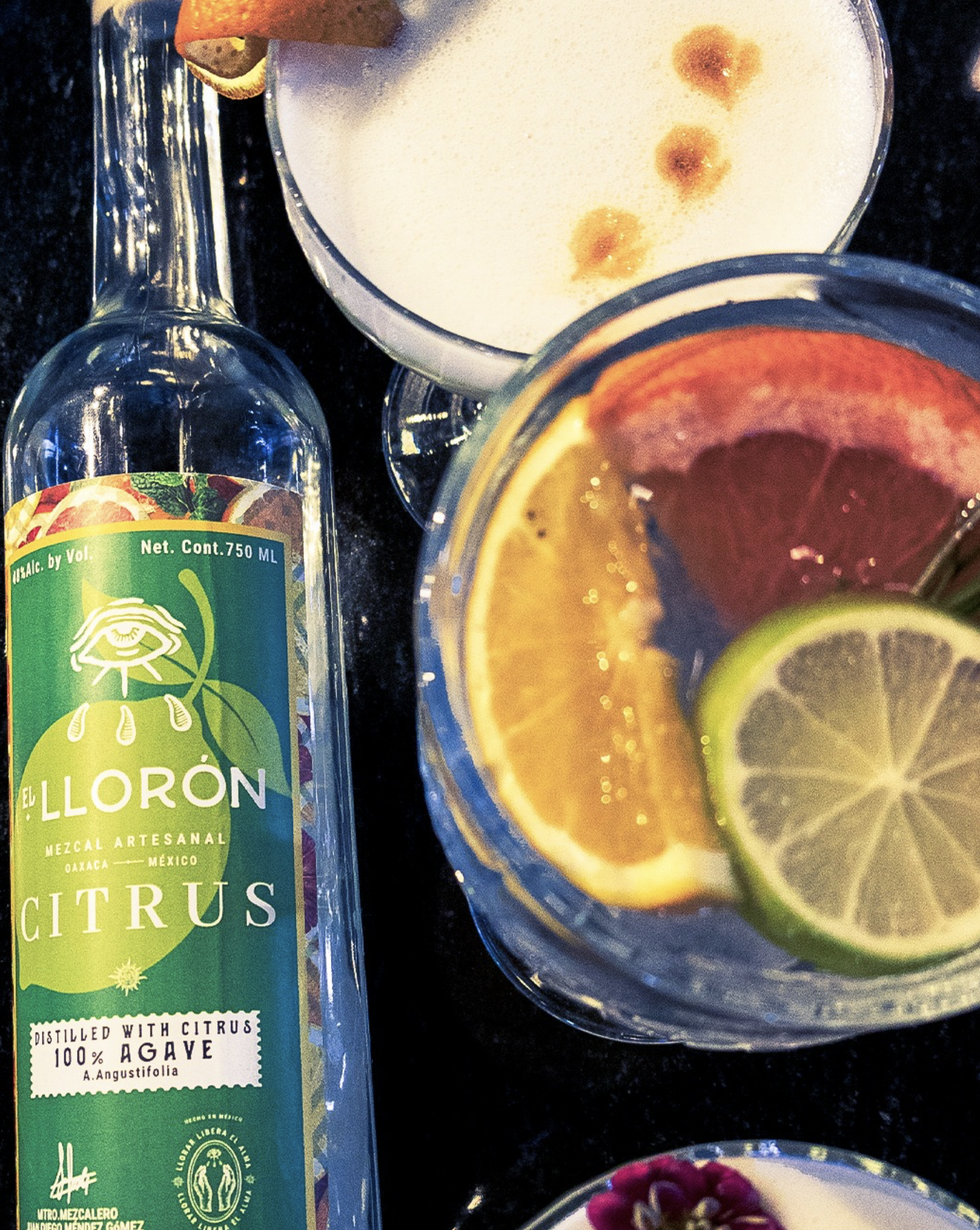 Botellas y vasos con diversas bebidas y rodajas de limón, naranja y otros ingredientes para preparar cócteles en un ambiente oscuro.