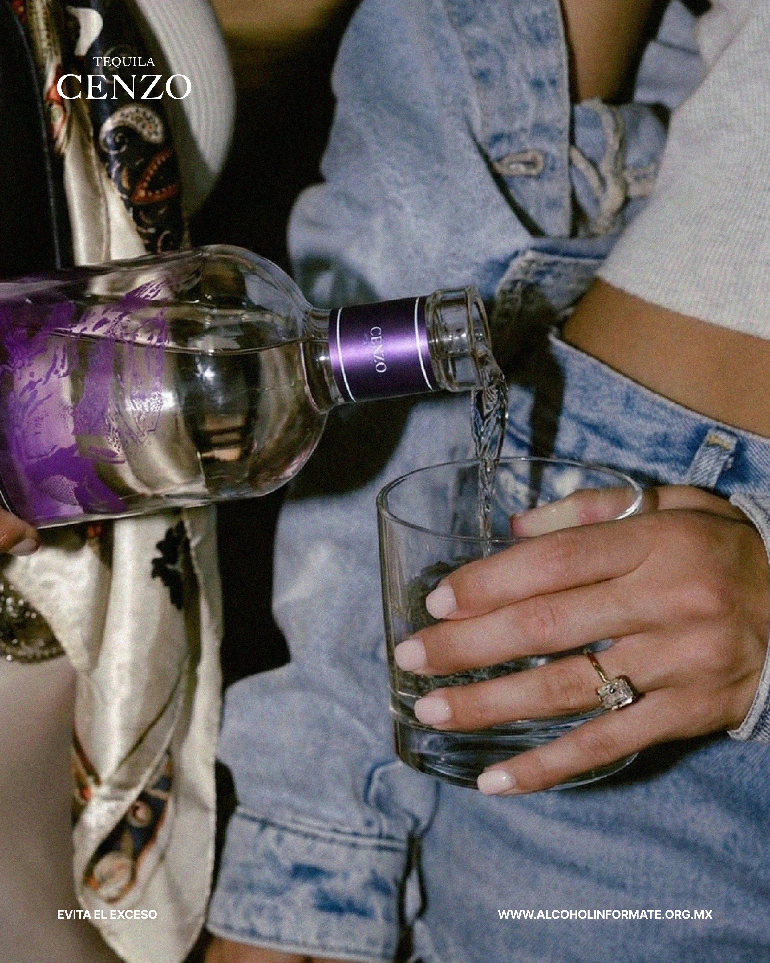 Una persona está sirviendo agua de una botella de tequila Cenzo en un vaso. La persona lleva un anillo y tiene las uñas pintadas. La persona viste un pantalón vaquero y una blusa de manga larga.