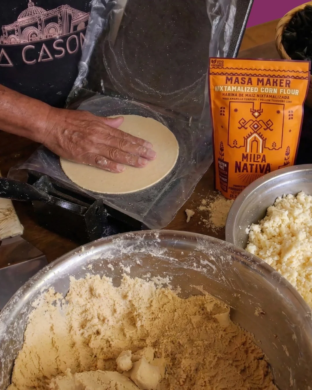 Persona formando una tortilla en una superficie con harina y maíz en grano, rodeada de ingredientes y utensilios para preparar masa de maíz.