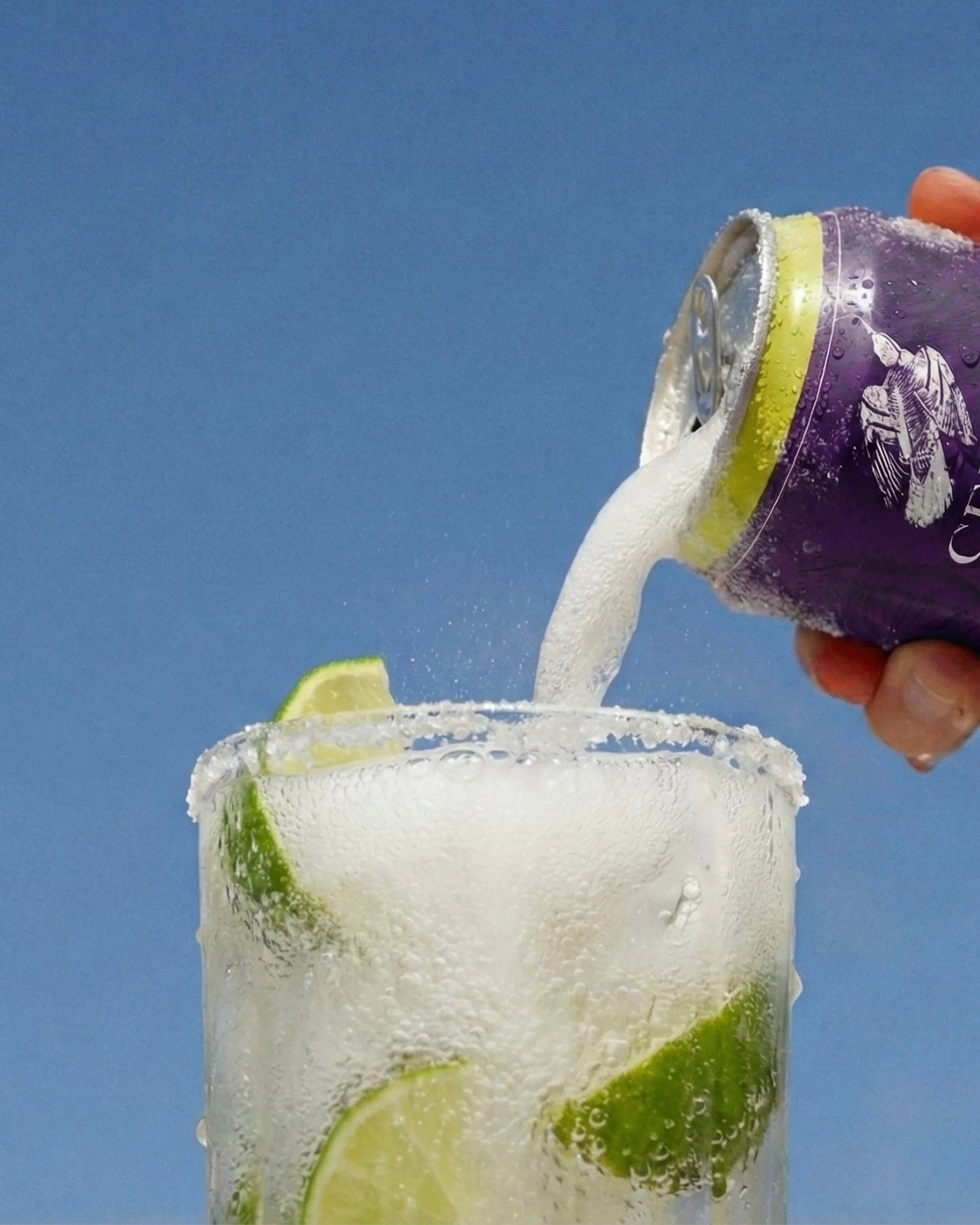 Una mano vierte refresco de lima-limón en un vaso con hielo y rodajas de limón y lima, con fondo azul.