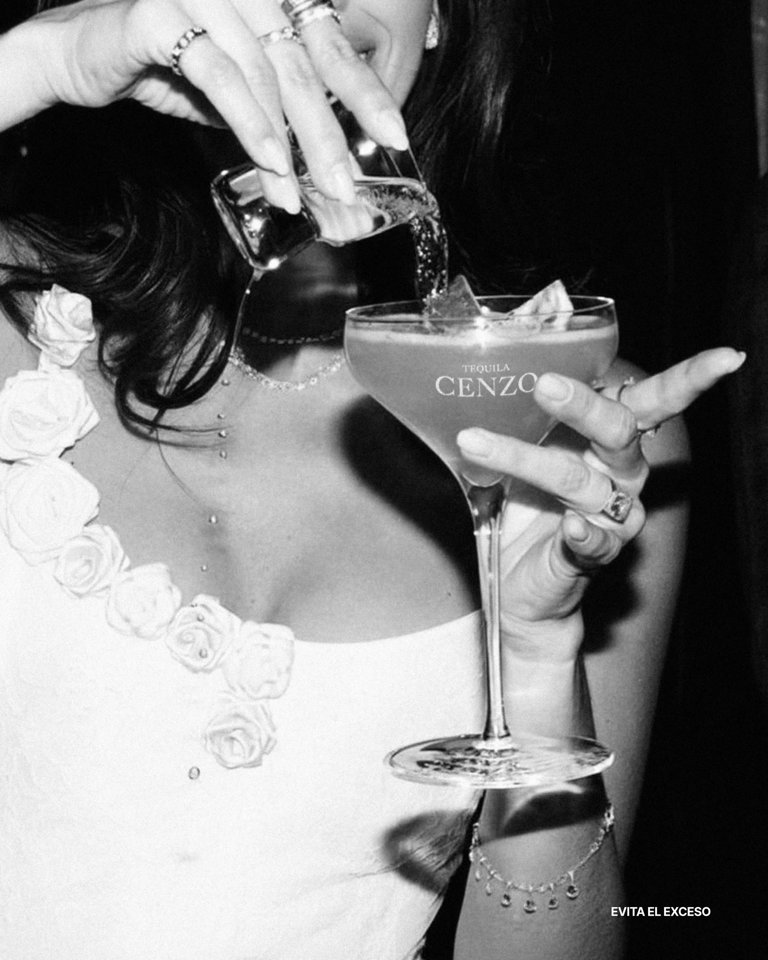 Una mujer con vestido blanco y accesorios sostenien un cóctel y sirve un licor en un vaso. Tiene uñas largas y usa varias joyas.