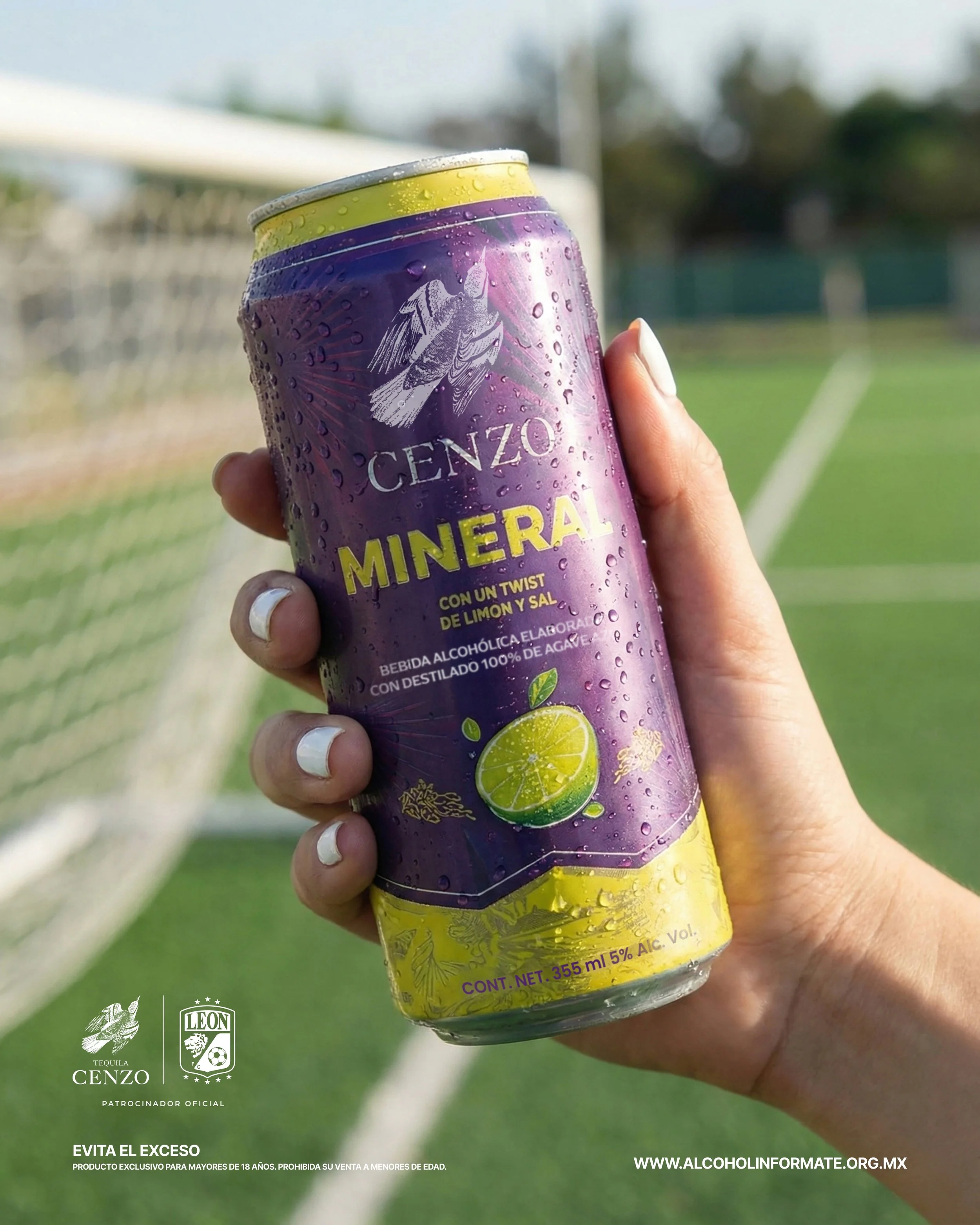 Lata de bebida alcohólica Cenzó Mineral con sabor a limón y sal, con gotas de agua en la superficie, siendo sostenida por una mano en un campo de fútbol durante el día.