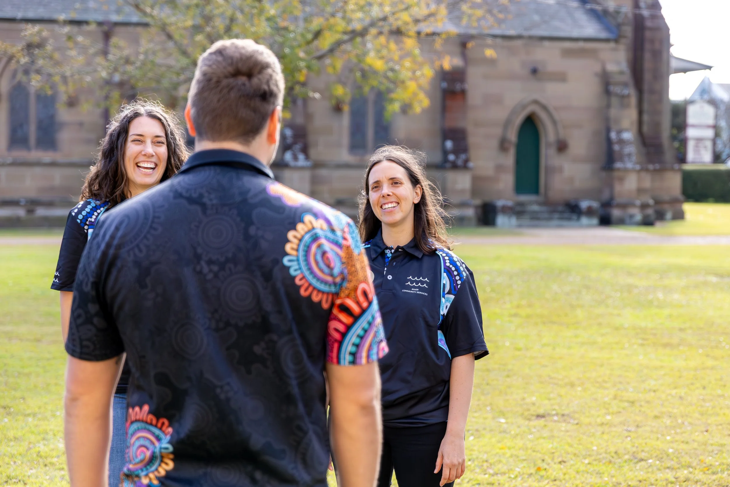 Three people, two women and one man, smiling and talking outdoors on a grassy area in front of a stone building, with trees in the background.