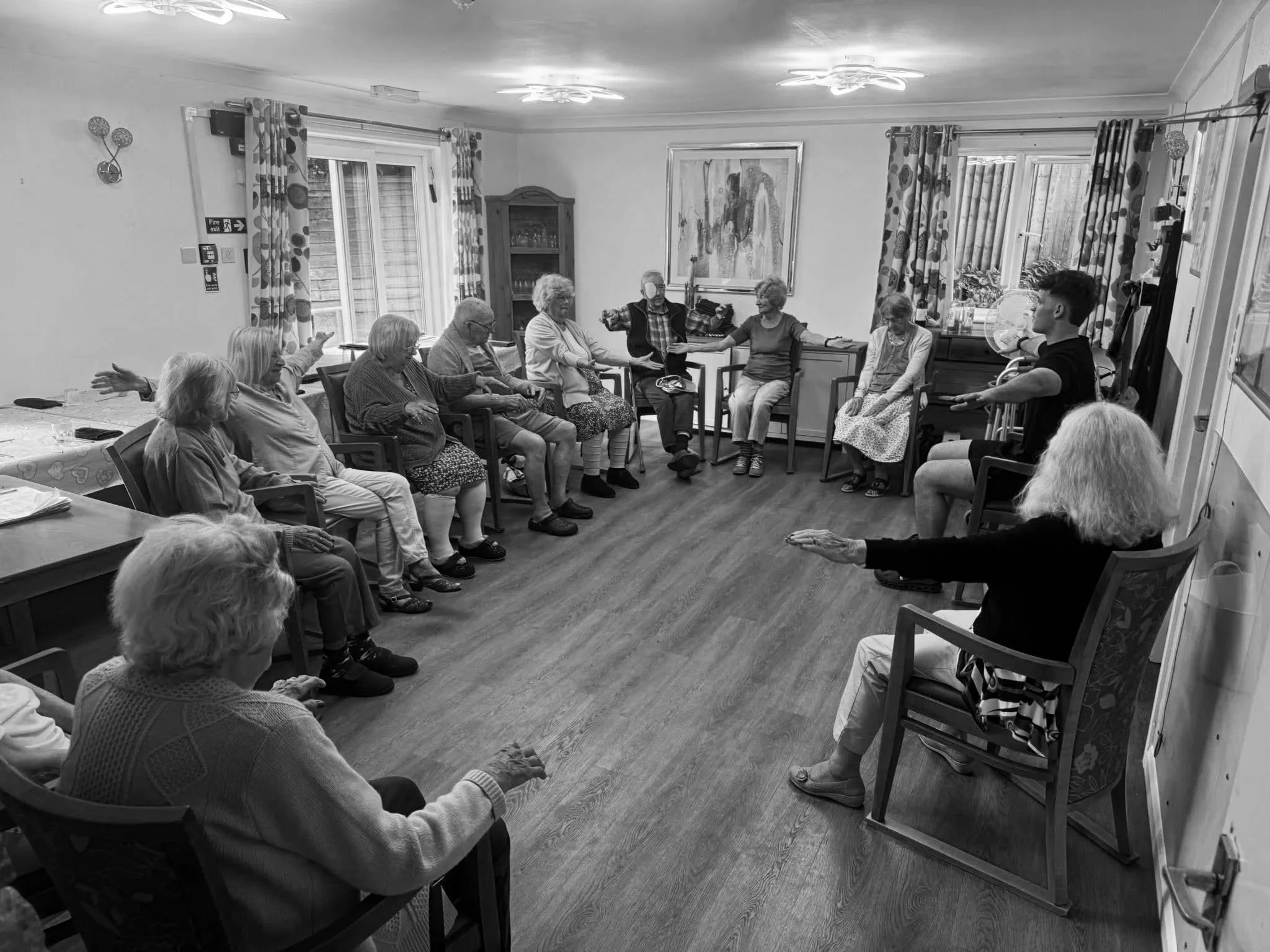 A group of elderly people participating in a seated exercise session in a room with wood flooring, decorated with curtains, a painting, and windows.