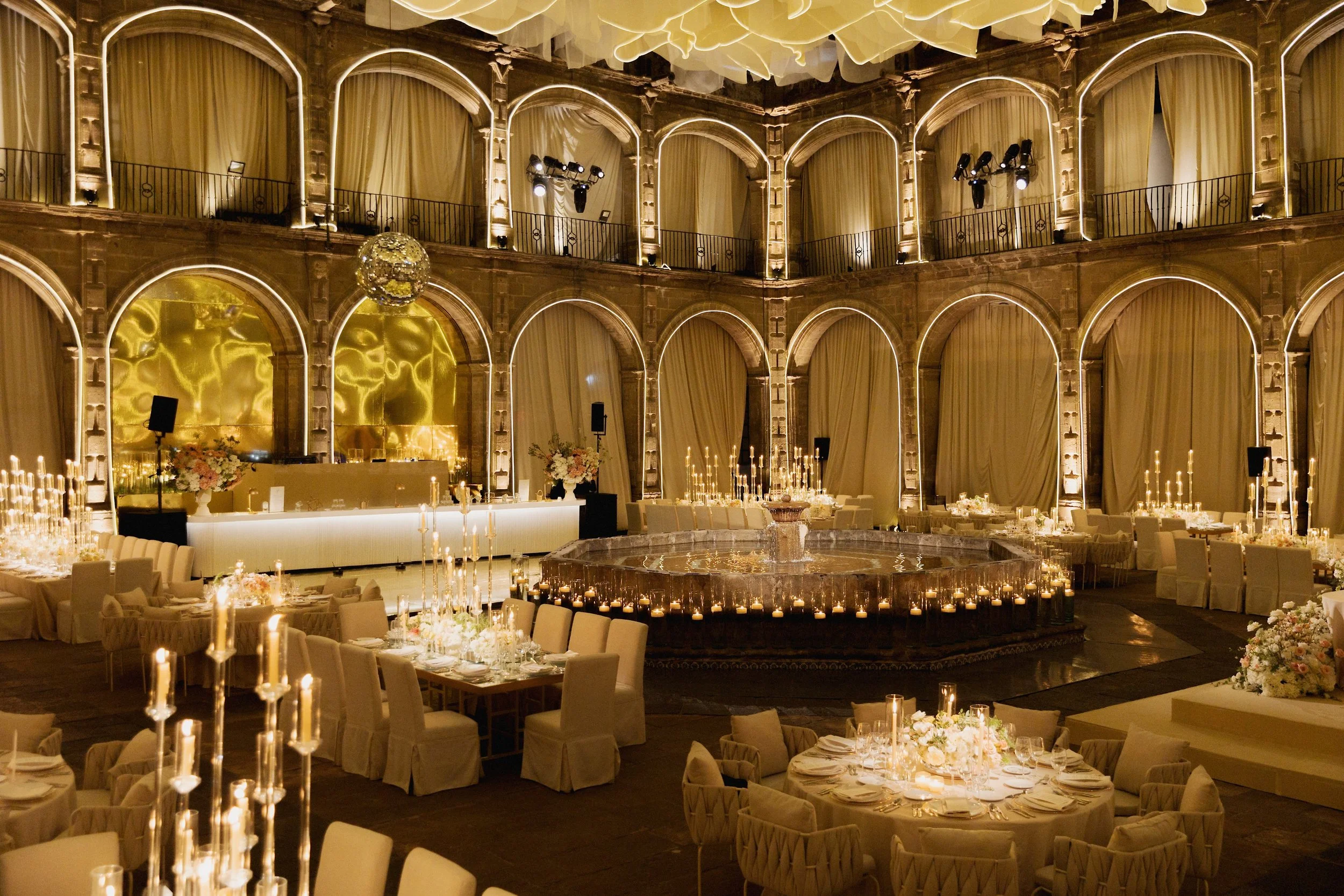 Elegant banquet hall decorated for a wedding reception with white draped tables, floral centerpieces, candelabras, and a dance floor, illuminated in warm lighting.