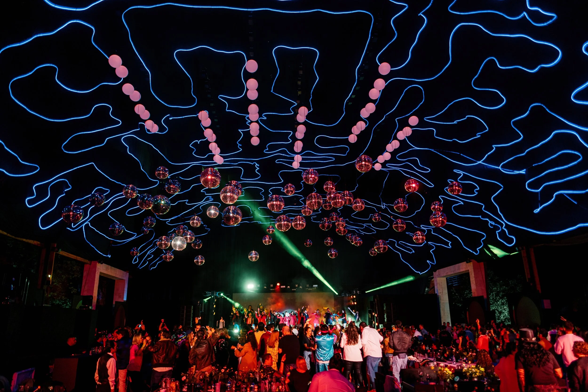 Elegant indoor event space decorated with abundant white and cream floral arrangements hanging from the ceiling and arranged on tables, illuminated by warm candlelight, with a stage or dance floor at the center.