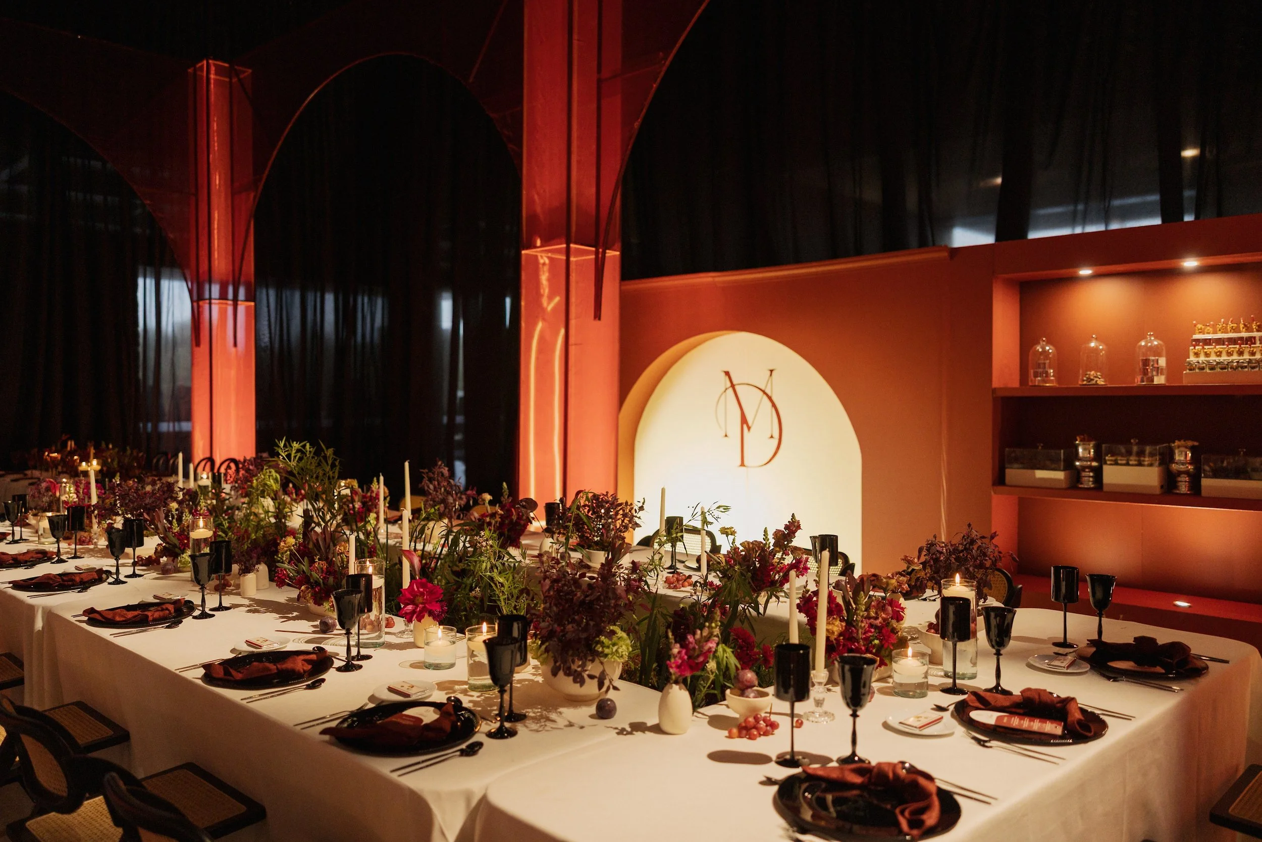 Elegant dining table set for an event with a long white tablecloth, black and copper glasses, floral centerpieces, and lit candles, in a dimly lit room with dark curtains and a lit wall with a monogram logo.