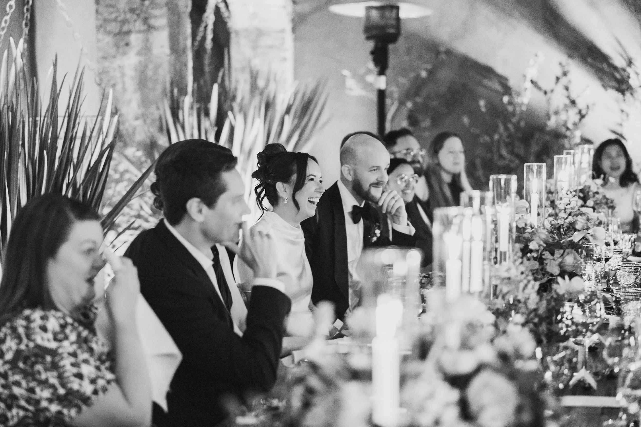 Close-up of an Indian bride and groom holding hands during a wedding ceremony. The bride is dressed in a red and gold traditional outfit with henna on her hand, wearing bangles, and a flower garland. The groom is in a cream-colored embroidered outfit with a flower garland.