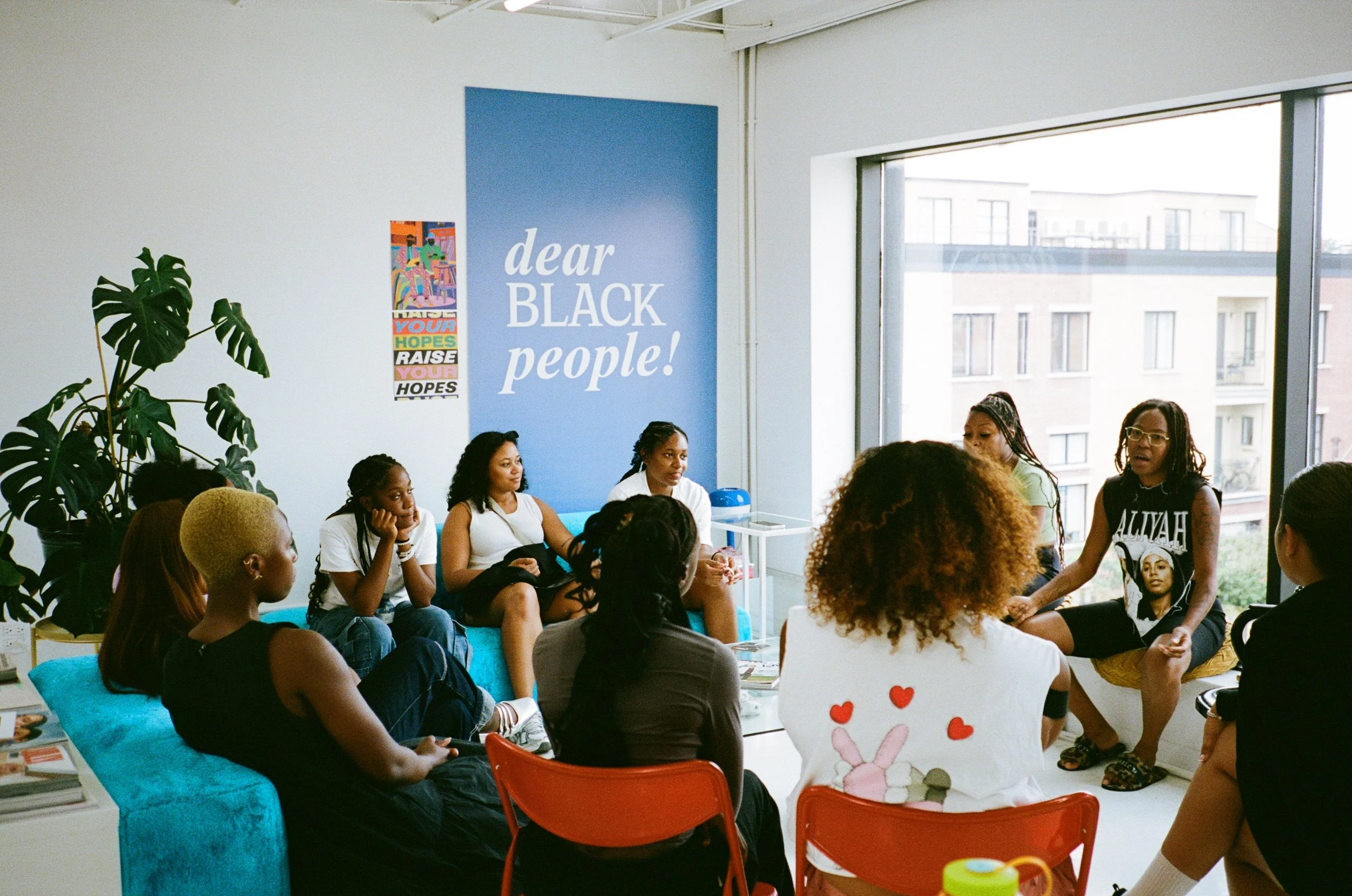Groupe de jeunes femmes, principalement noires, assises dans un salon moderne avec une grande baie vitrée. Certaines parlent, d'autres écoutent, dans une ambiance de réunion ou atelier. Mur bleu avec message « dear BLACK people! » et affiche colorée sur le mur.
