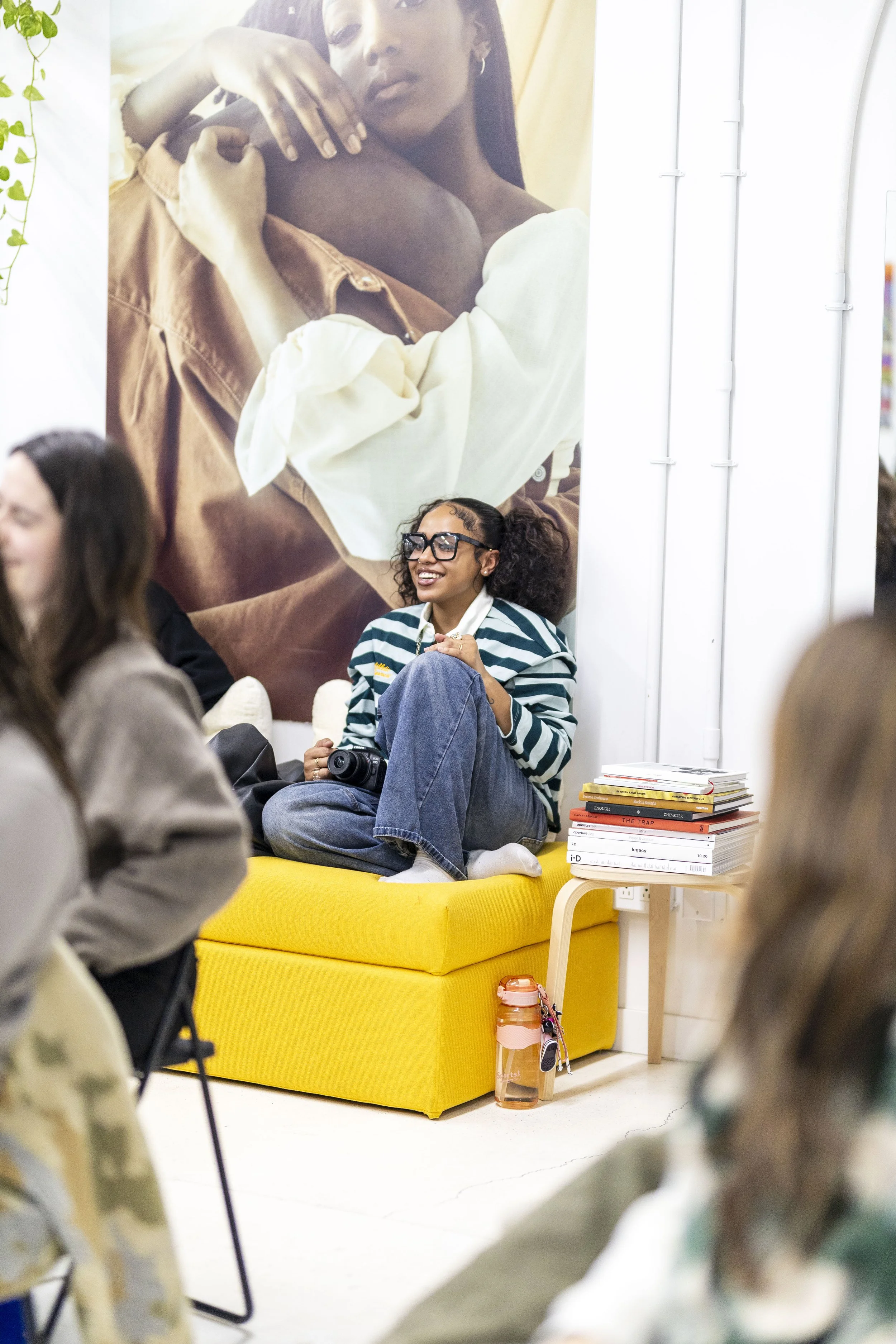 Une femme souriante assise sur un canapé jaune, portant des lunettes épaisses et un pull à rayures, entourée de plusieurs personnes dans une ambiance décontractée, avec une grande affiche d'une autre personne en arrière-plan.
