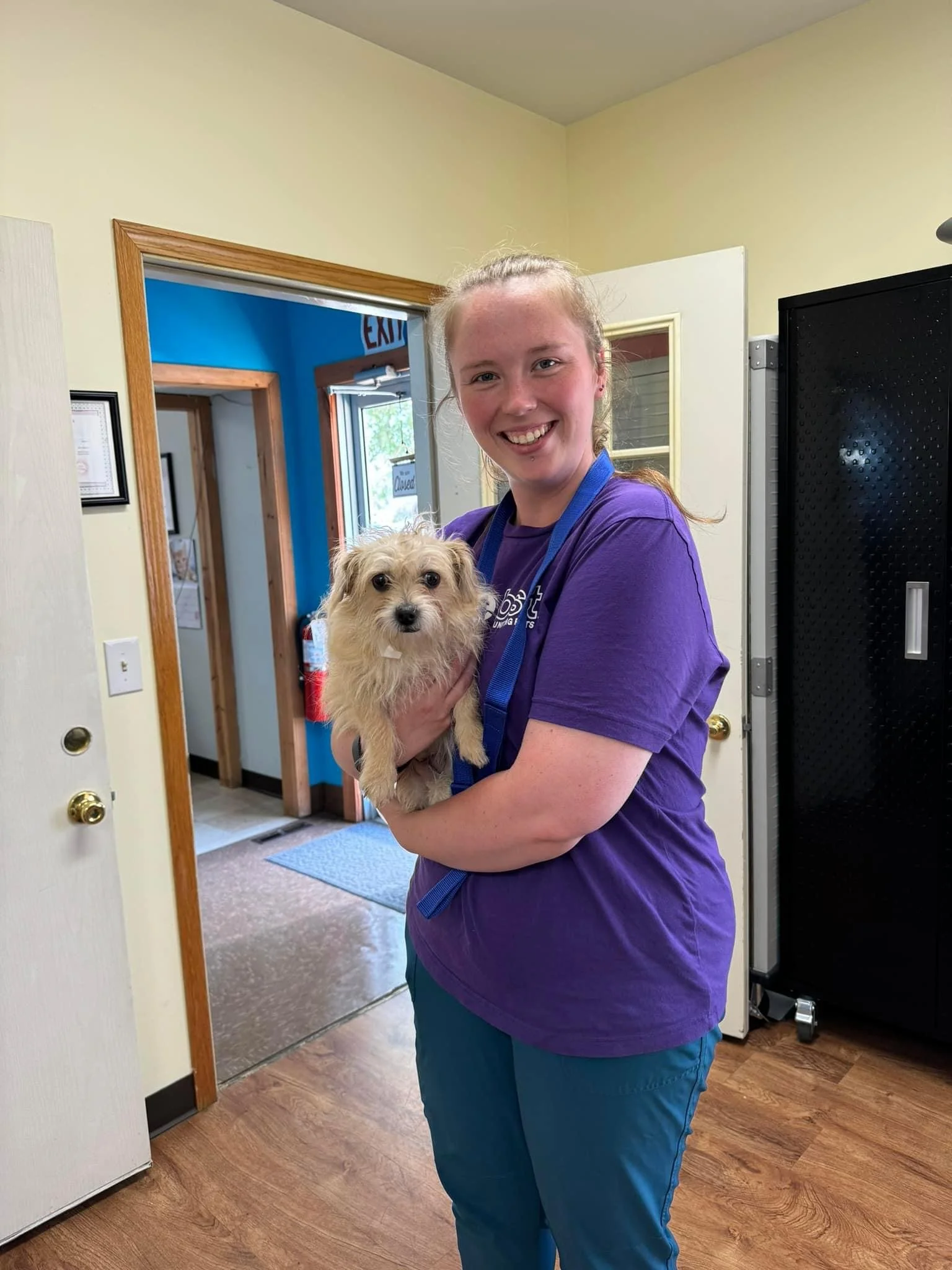A smiling young woman holding a small, fluffy dog indoors getting love before vaccines in a vet clinic called Spay Arkansas.