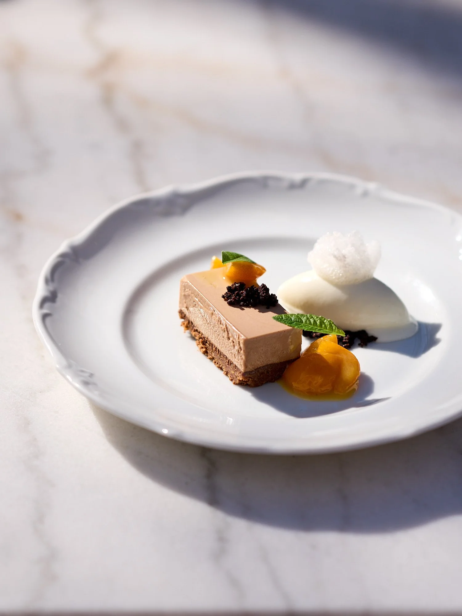 Ein Teller mit einem Stück Schokoladenkuchen, weißer Creme, Früchten und Minzblättern, auf einem weißen Marmortisch.