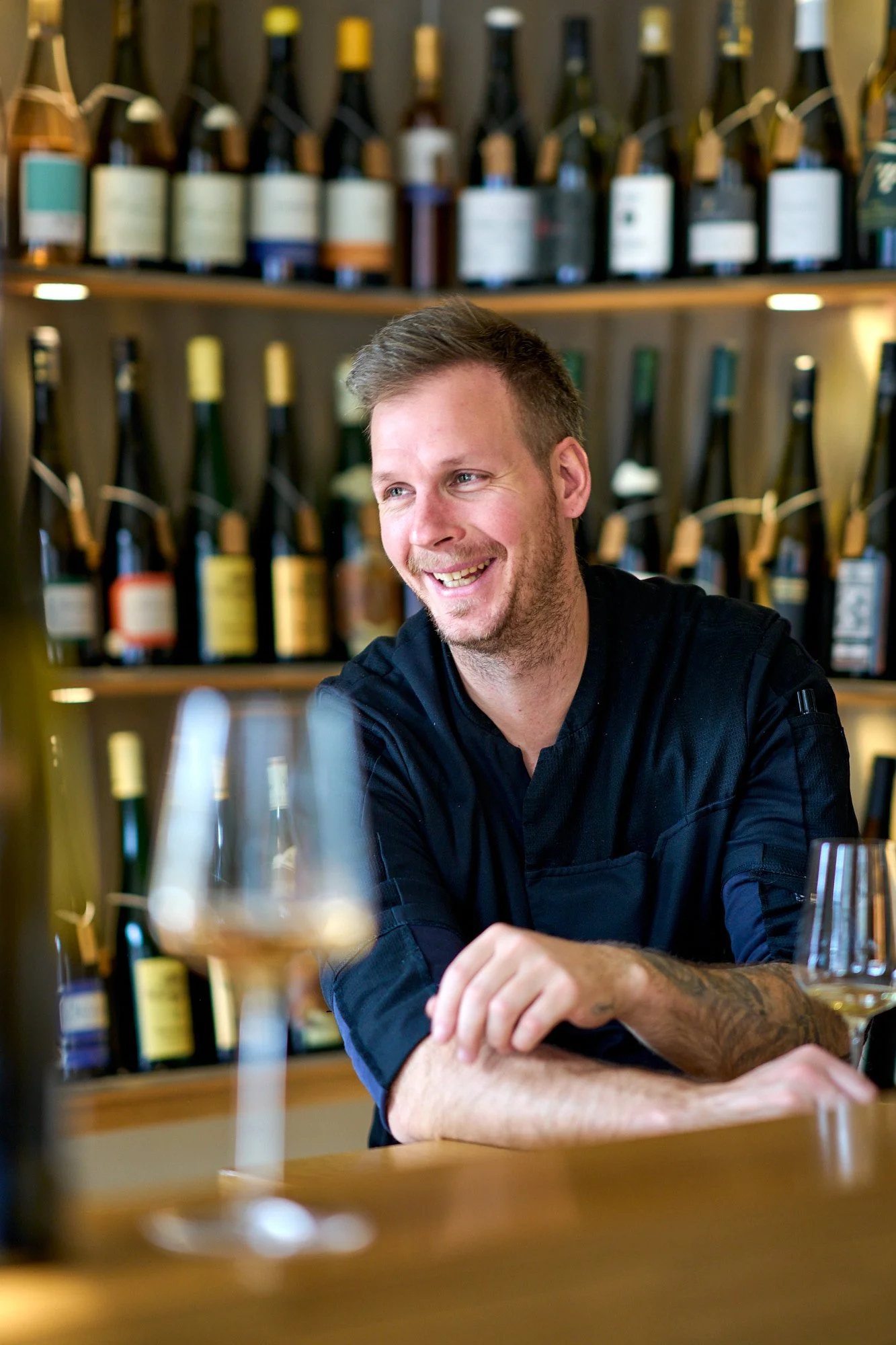 Ein lachender Mann sitzt in einem Restaurant mit Weinen im Hintergrund, darunter auch Weinflaschen an der Wand.