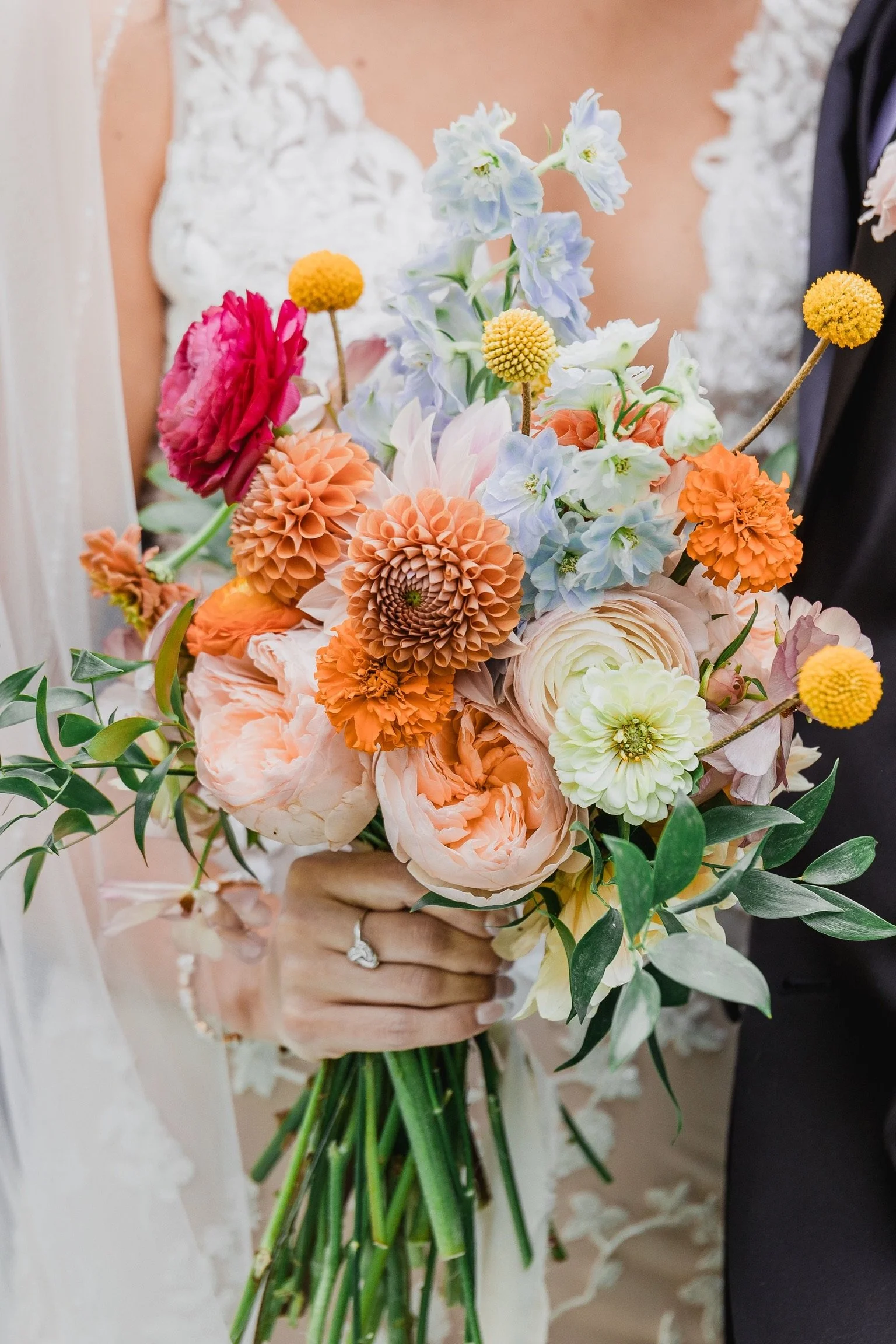 A bride and groom holding a vibrant bouquet of flowers that includes peach, pink, and white blossoms with green foliage, with the bride wearing a white lace dress and the groom in a black suit.