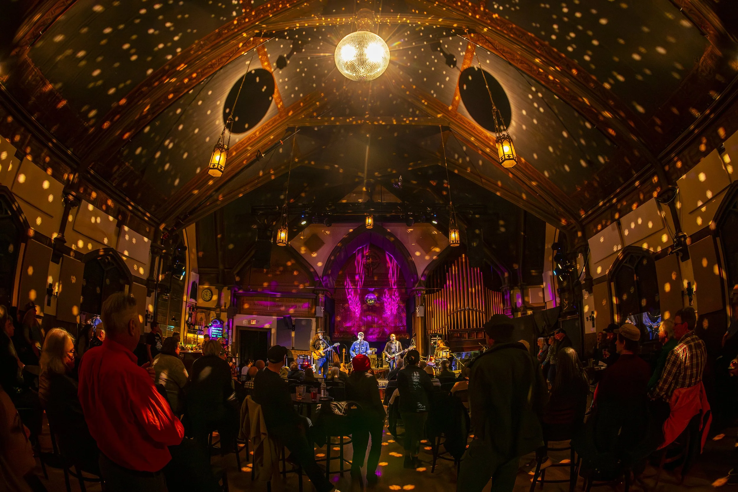 Indoor concert venue with a live band performing on stage, audience standing and seated, colorful lighting, and a disco ball reflecting lights on the ceiling.
