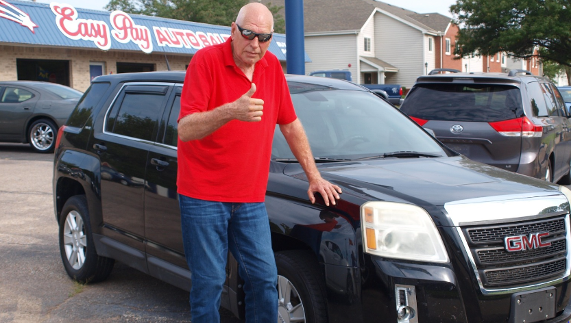 A man in a red shirt and jeans giving a thumbs-up next to a black GMC SUV at Easy Pay Auto Sales.