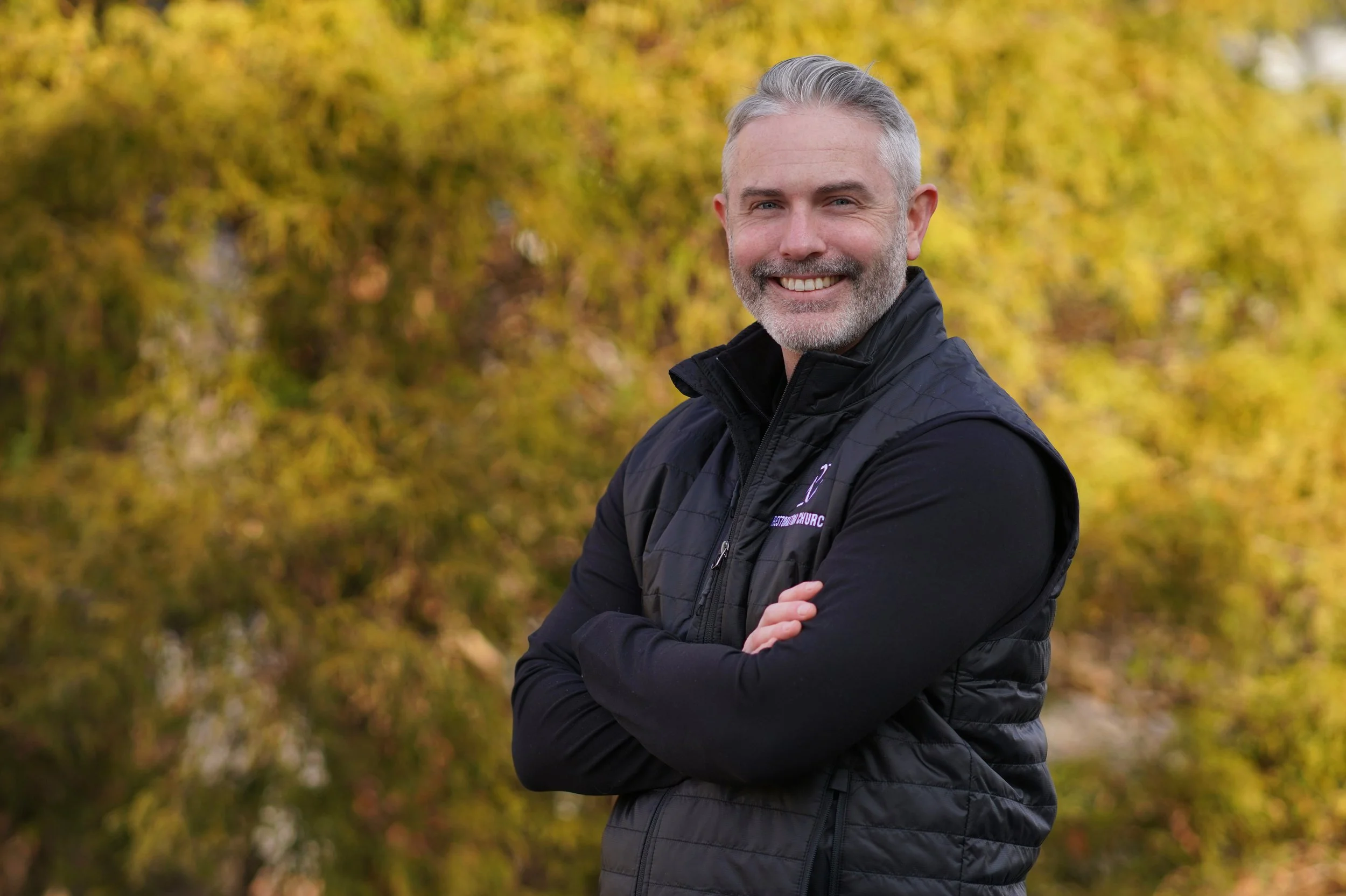 A smiling man with gray hair and a beard, wearing a black jacket, standing outdoors in front of autumn foliage.