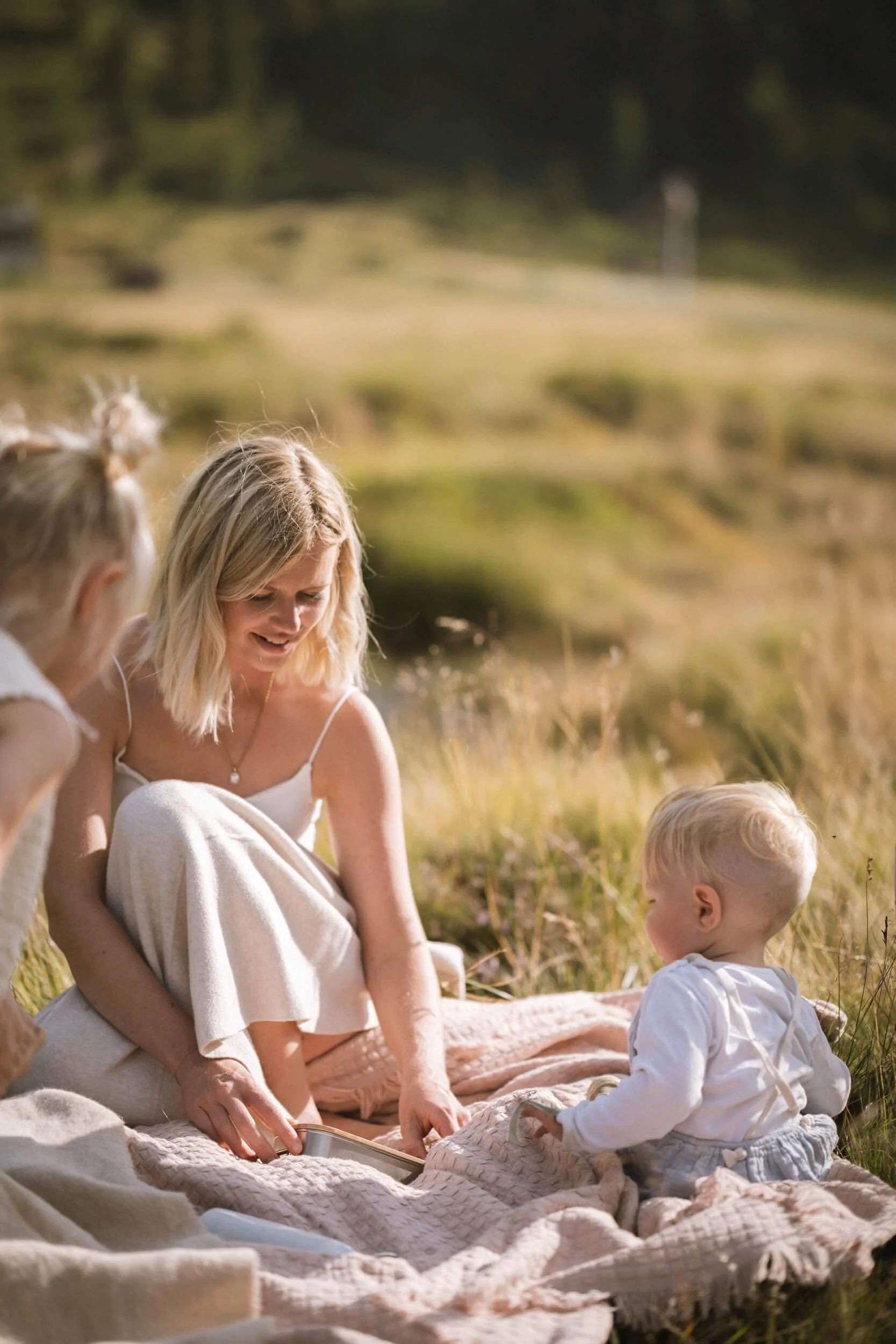 Eine Frau und zwei Kinder sitzen auf einer Decke im Freien, umgeben von Natur, während sie zusammen ein Buch anschauen.