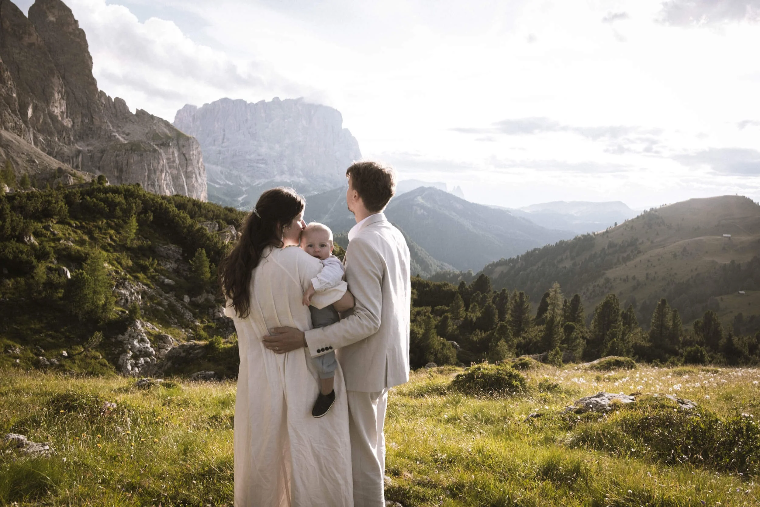 Eine Familie mit einem Kind in einer grünen Berglandschaft, umgeben von Bergen und Wäldern bei Sonnenuntergang.