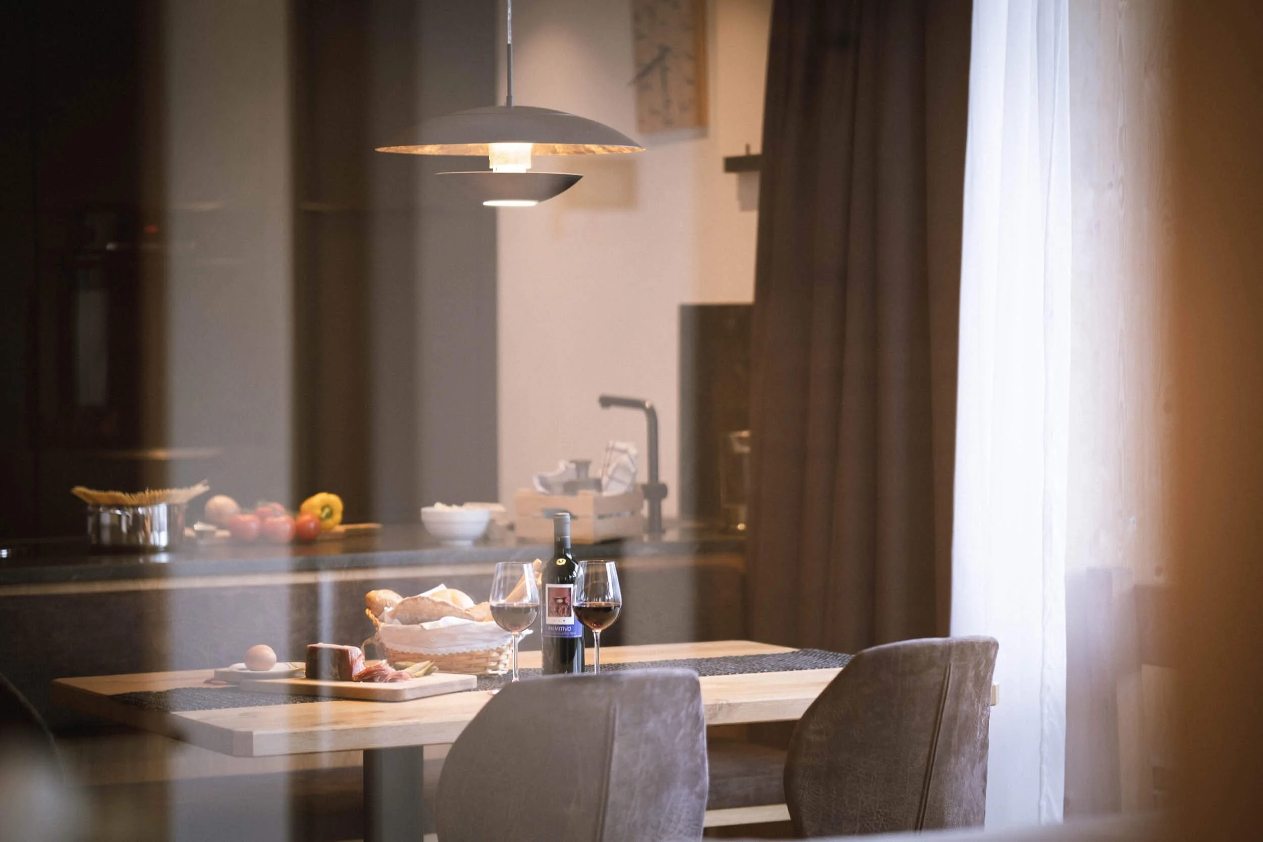 Ein gemütlich eingedeckter Esstisch mit Wein, Brot und Käse in einer hellen, warm beleuchteten Küche, sichtbar durch eine Fensterscheibe.