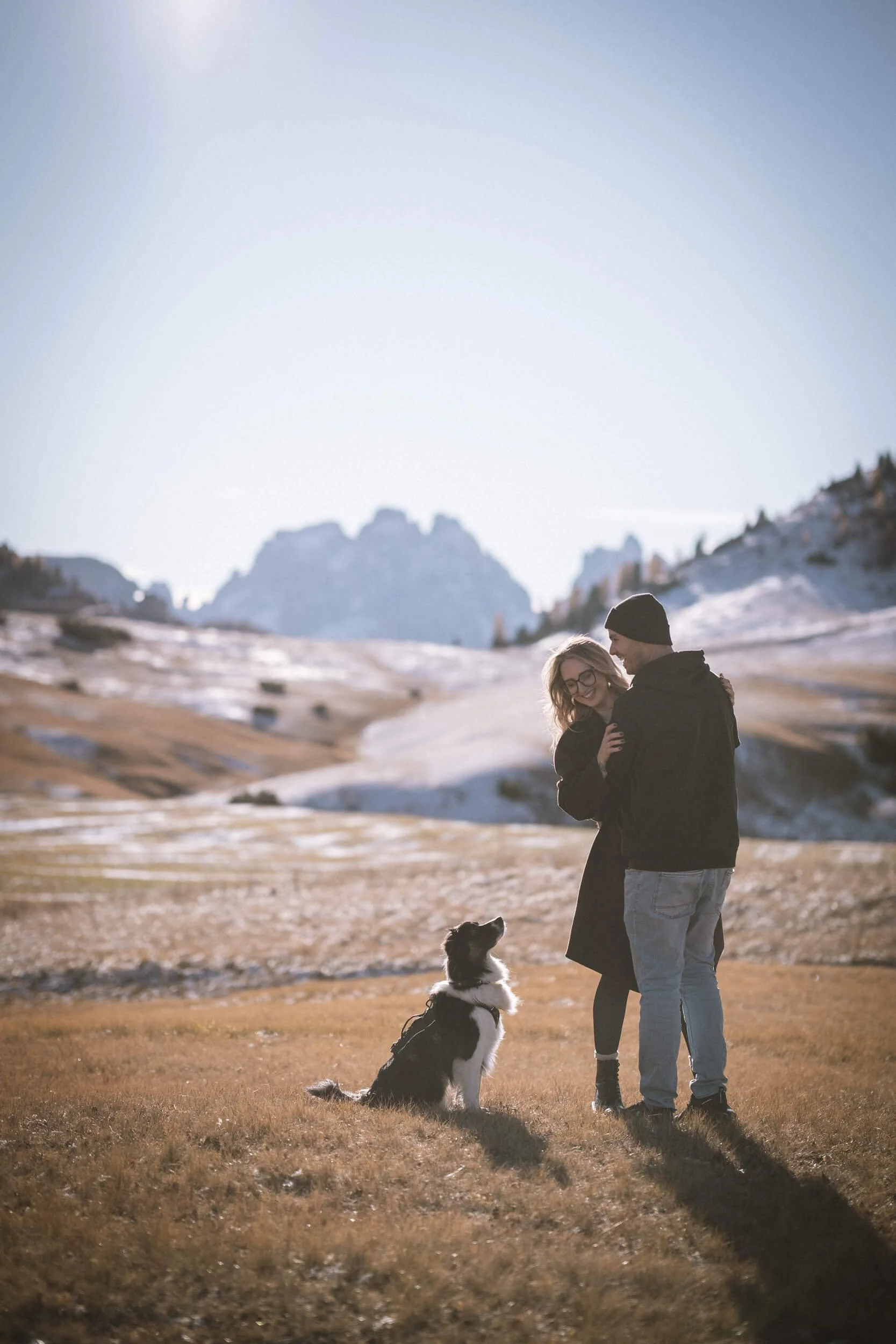 Ein Paar mit Hund in einer winterlichen bergigen Landschaft, Sonne im Hintergrund.
