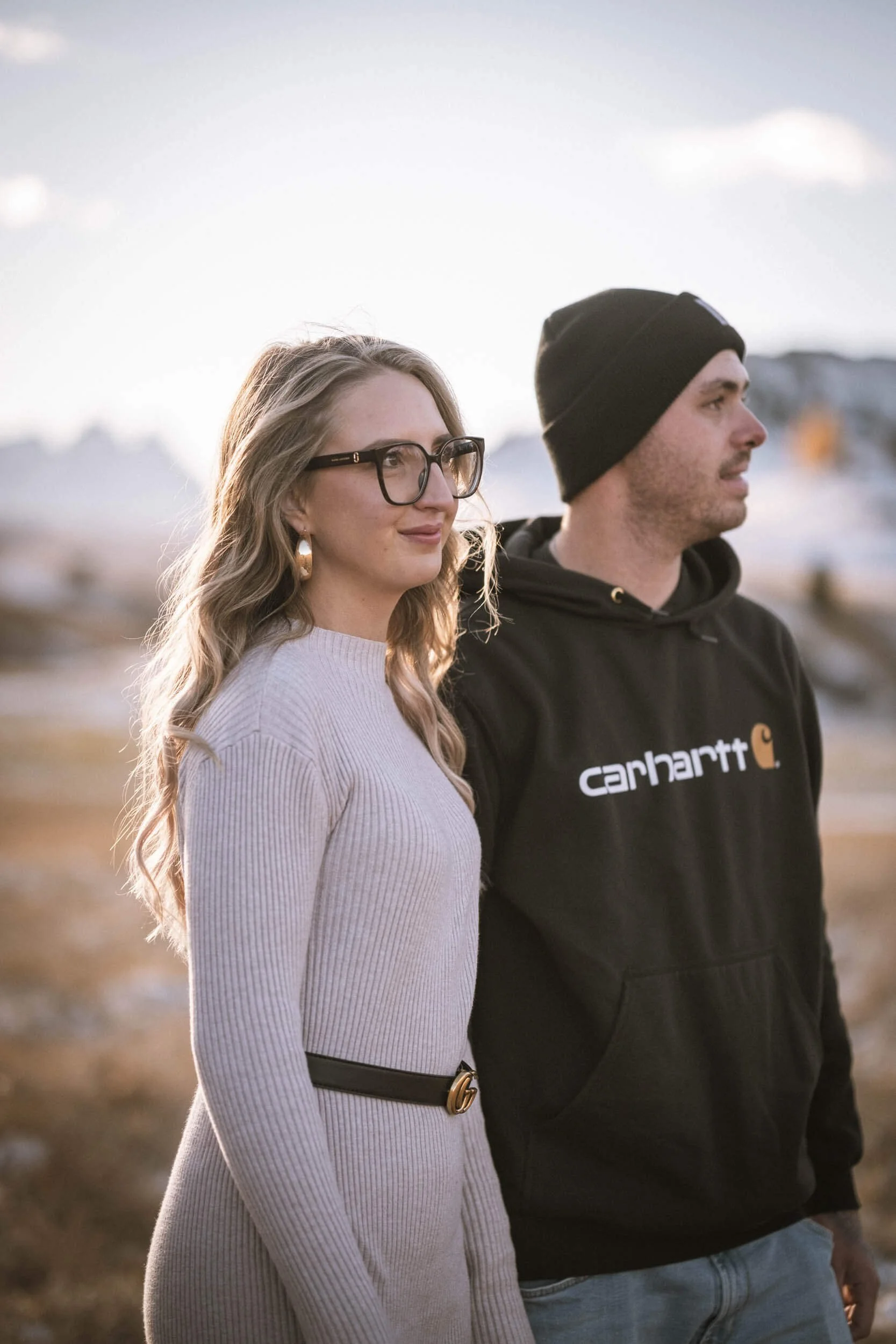 Ein Mann und eine Frau stehen nebeneinander im Freien bei Sonnenuntergang, im Hintergrund Berge und Himmel.