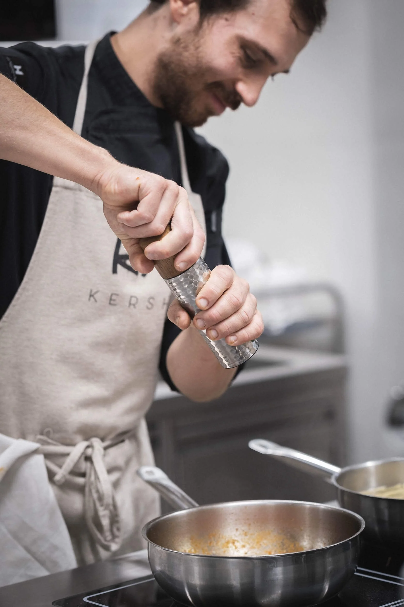 Ein Mann mit Brauen, dunklen Haaren und Bart, der in einer Küche Pfeffer auf eine Pfanne streut.