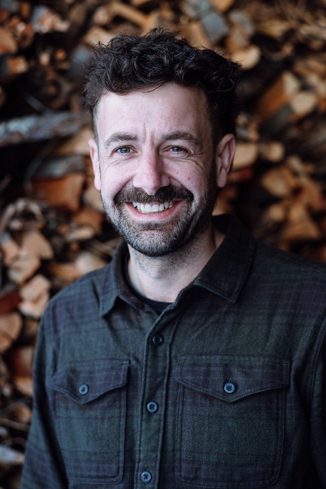 Ein lachender Mann mit dunklem, gewelltem Haar und Bart vor einem Stapel Holzscheite.